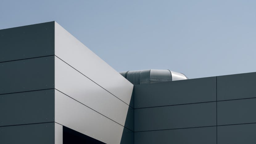 Modern building exterior with clean lines made of dark gray materials, viewed against a clear blue sky. A metallic ventilation duct is partially visible on the roof.