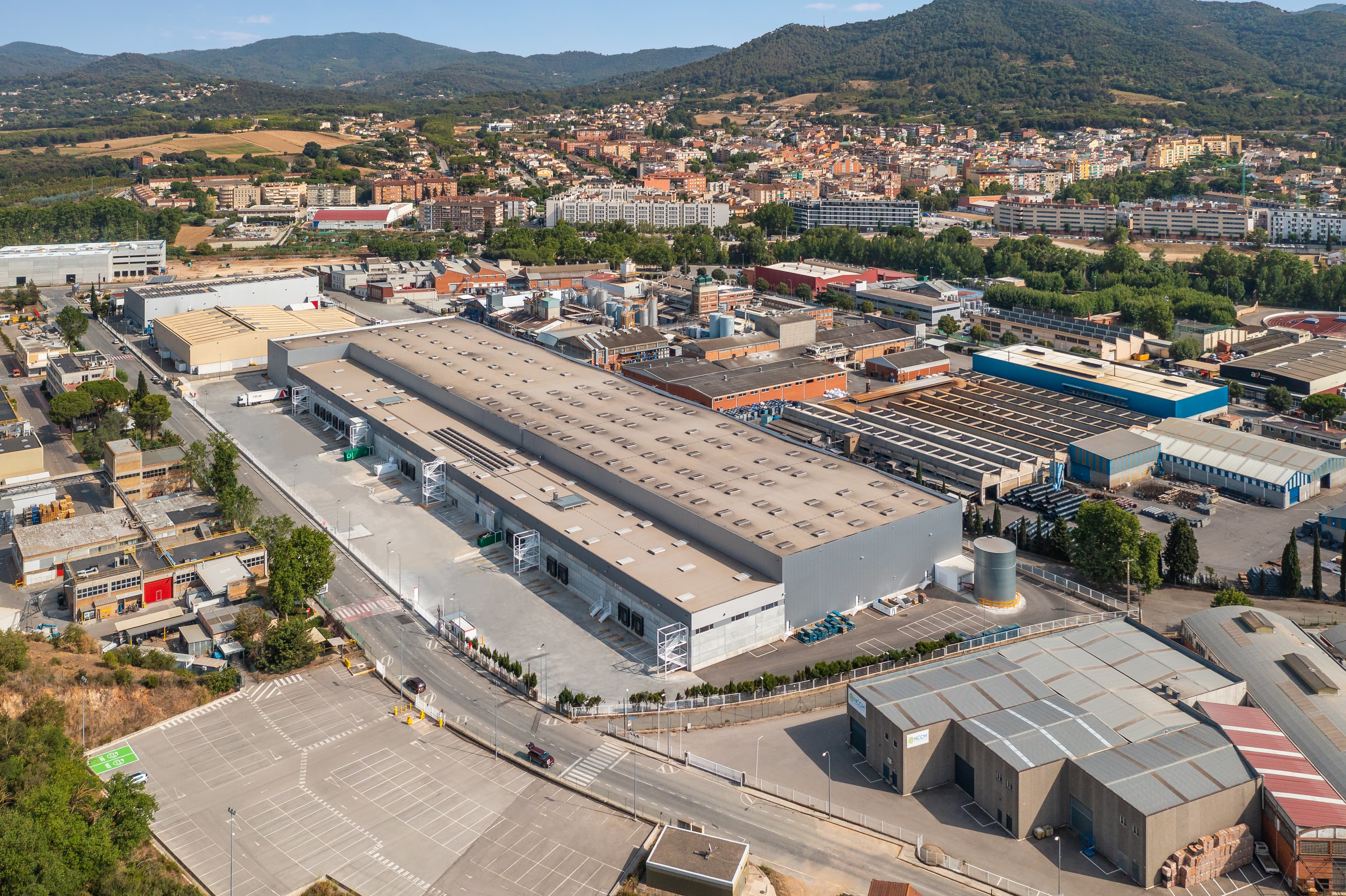 Vista aérea de una amplia instalación industrial rodeada de zonas de aparcamiento, con una ciudad y colinas al fondo.