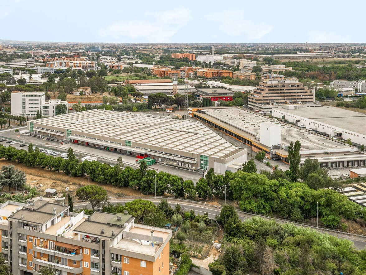 Vista aérea de un complejo industrial rodeado de vegetación y edificios, con un paisaje urbano al fondo bajo un cielo nublado.