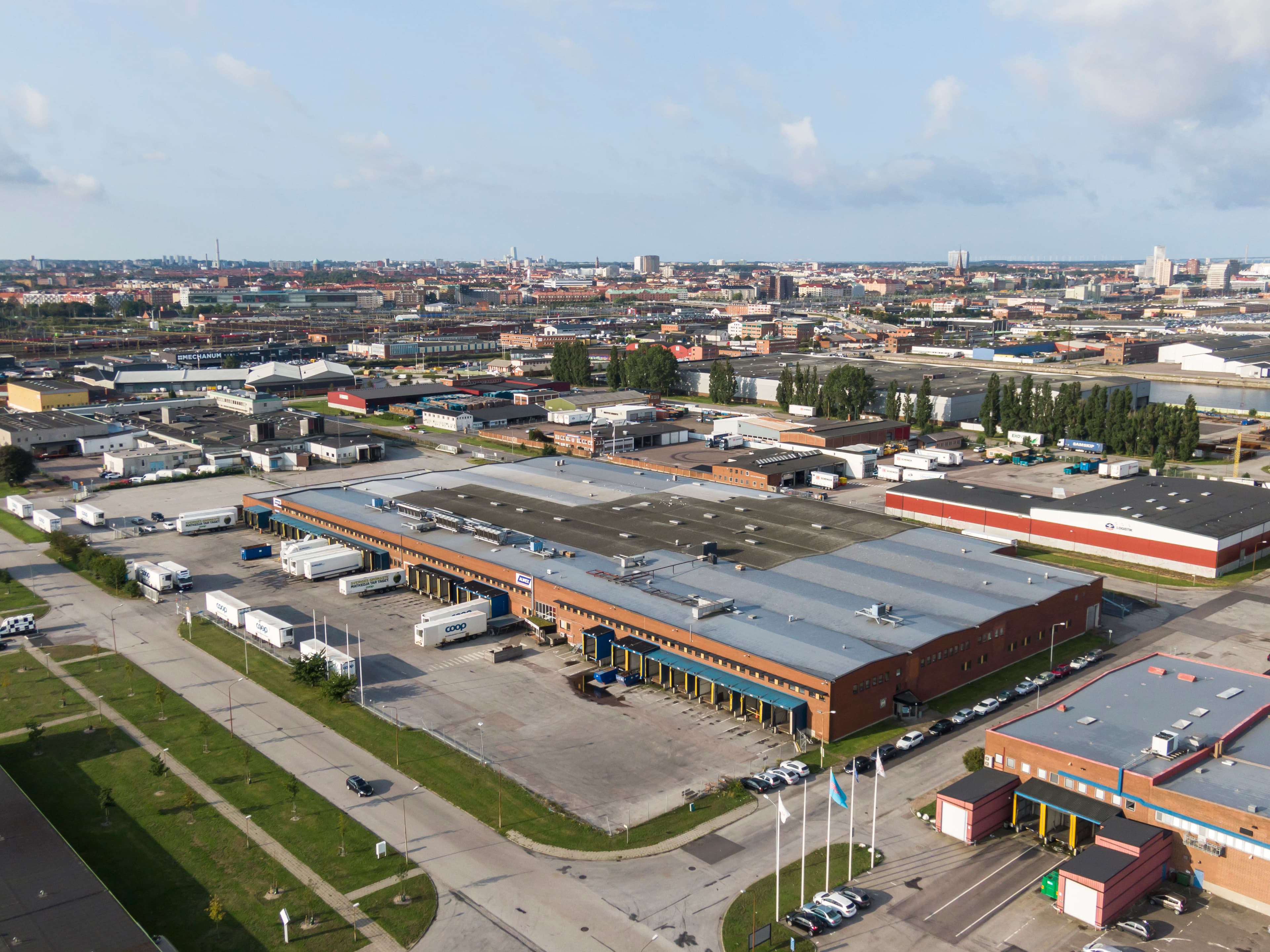 Vista aérea de una zona industrial con grandes almacenes, camiones y plazas de aparcamiento, rodeada de edificios urbanos bajo un cielo parcialmente nublado.