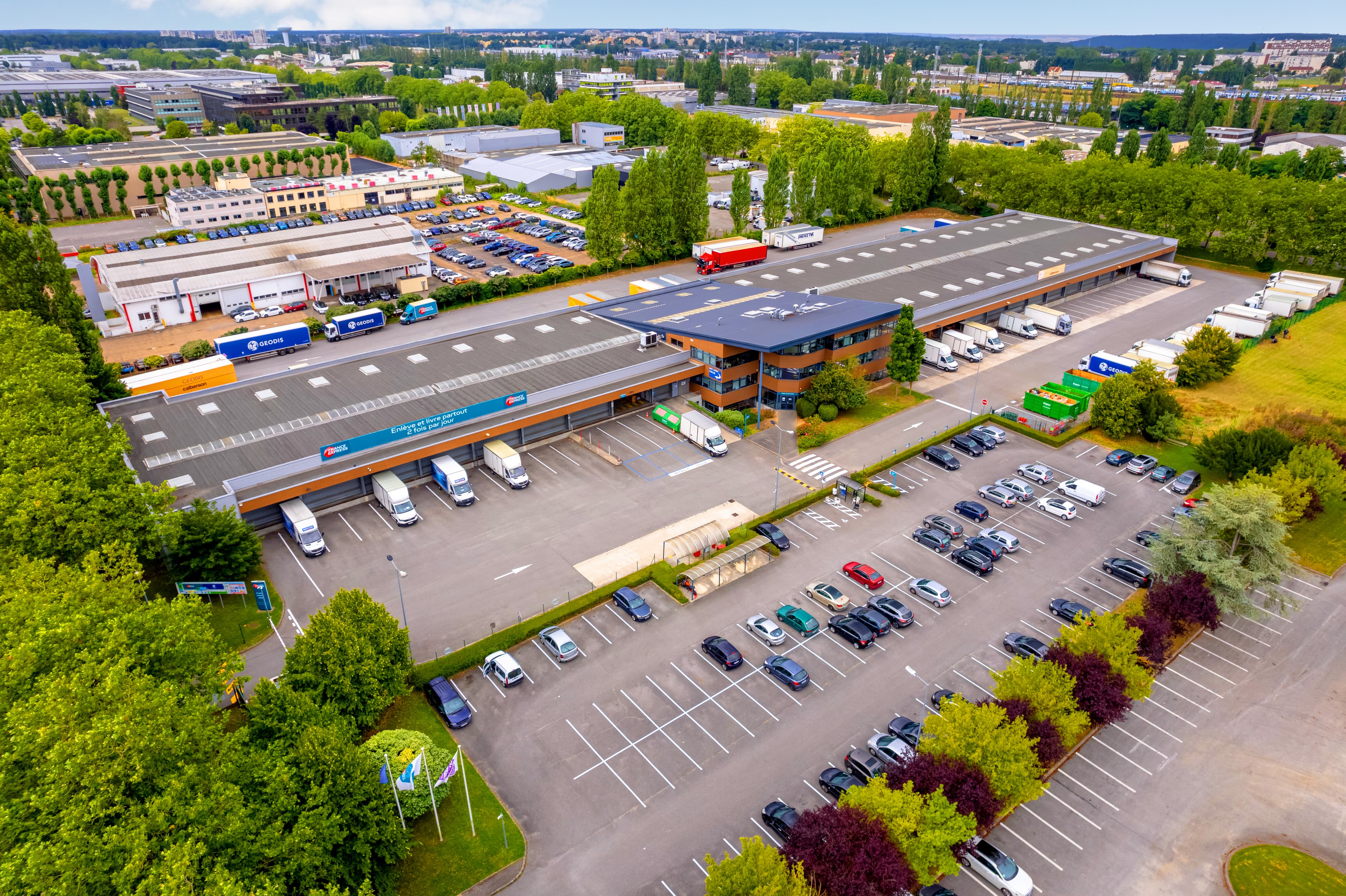 Vista aérea de un gran complejo de almacenes con muelles de carga, rodeado de camiones y automóviles estacionados, ubicado en una zona industrial verde.