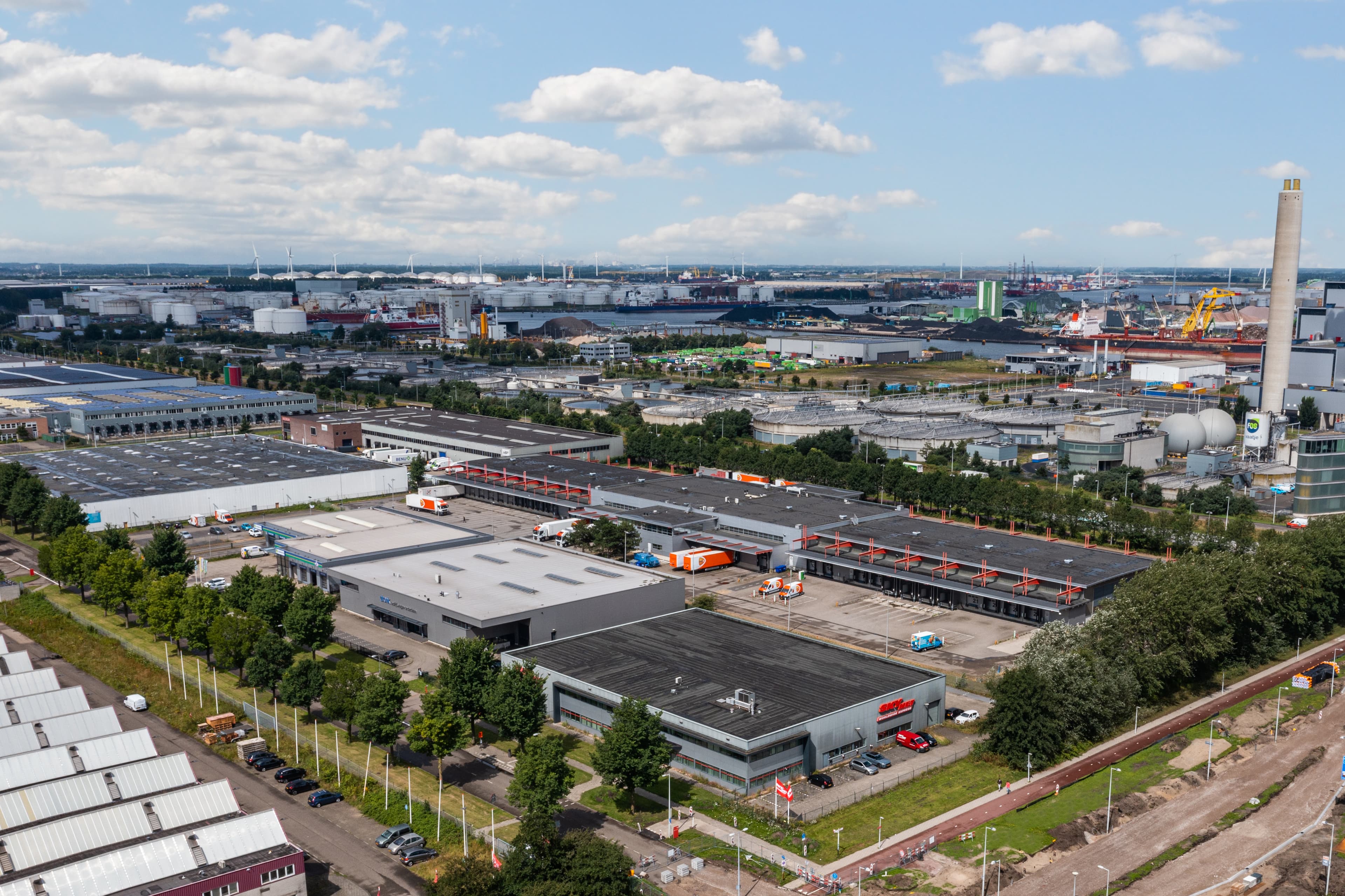 Luftaufnahme eines Industriegebiets mit Lagerhallen, Fabriken und Parkplätzen unter einem teilweise bewölkten Himmel.