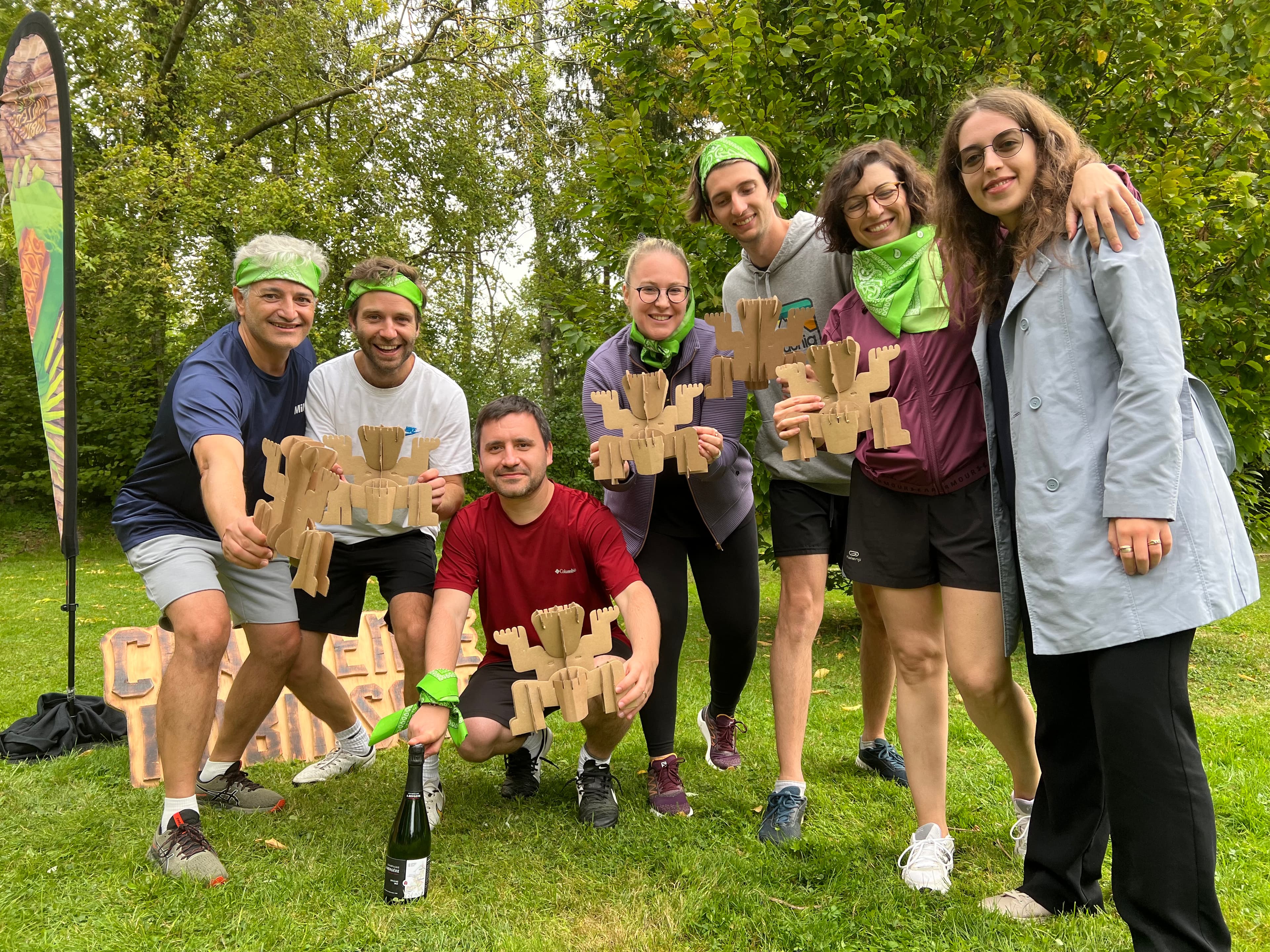 Een groep mensen buiten met houten trofeeën, sommigen dragen groene bandana's. Ze lachen en poseren voor de foto, staand op gras met bomen op de achtergrond.