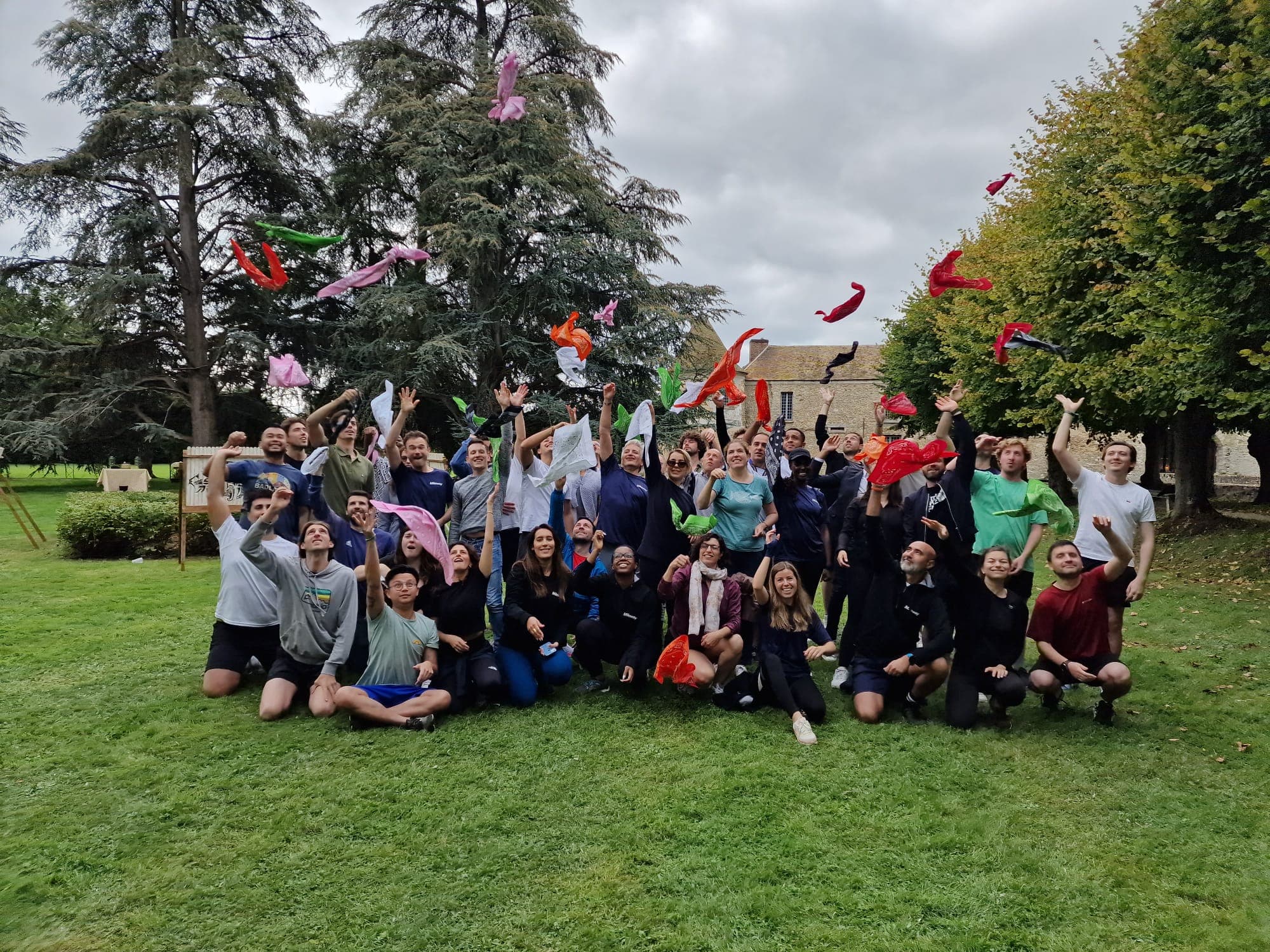 Een groep mensen buiten gooit kleurrijke sjaals in de lucht. Ze staan en zitten op het gras, met bomen op de achtergrond.