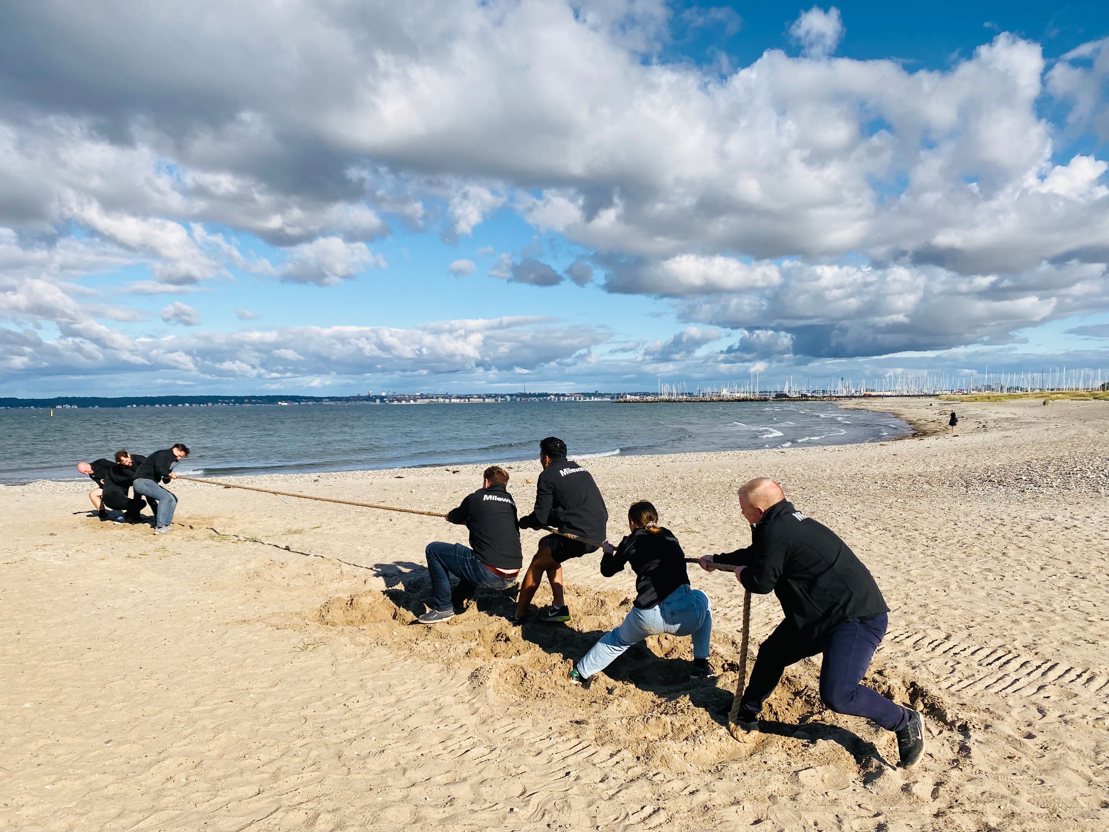 Een groep mensen is verwikkeld in een touwtrekwedstrijd op een zandstrand onder een gedeeltelijk bewolkte hemel.