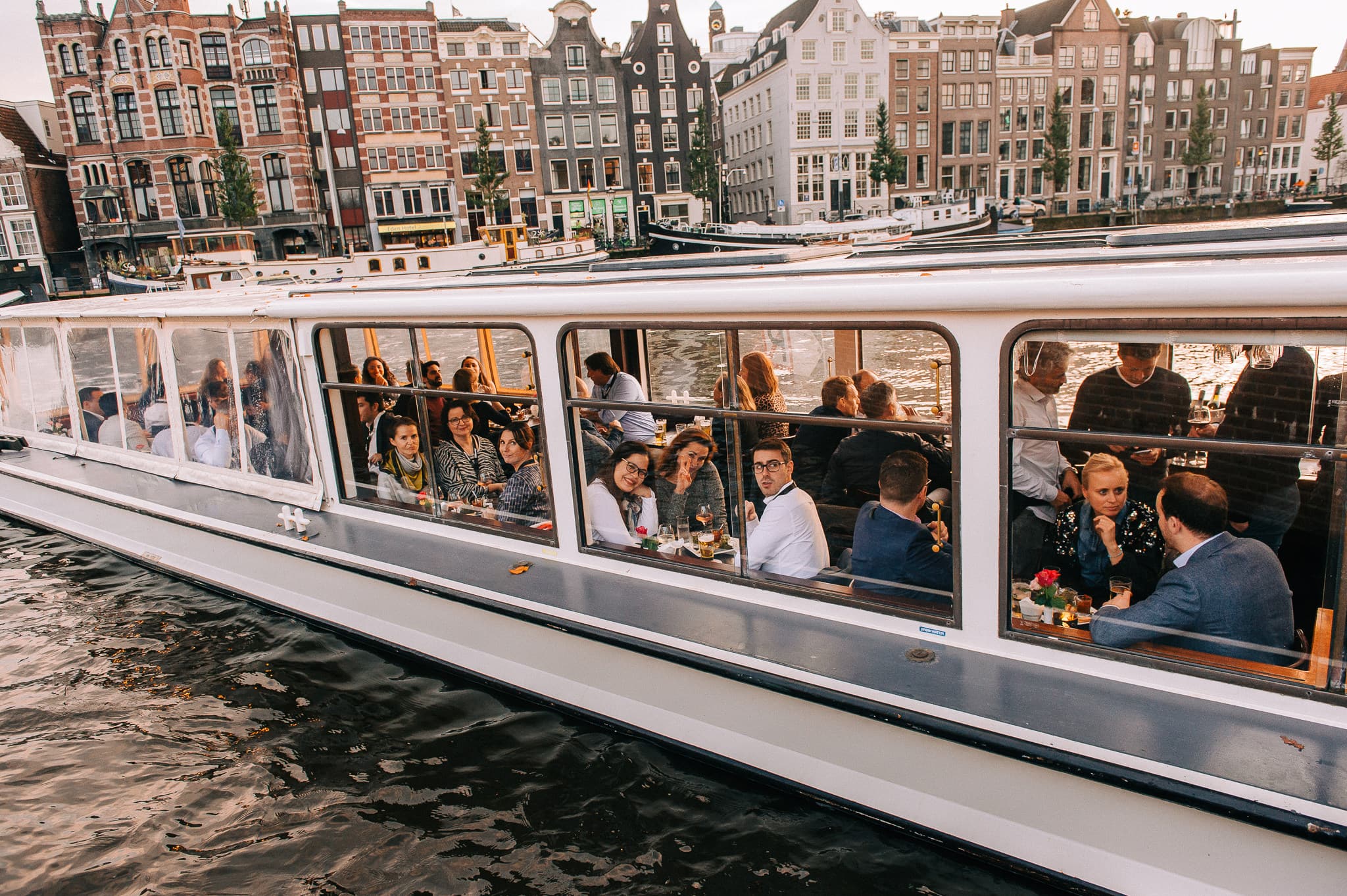 Een groep mensen eet in een kanaalboot met een glazen dak. De boot vaart door een kanaal met historische gebouwen op de achtergrond.