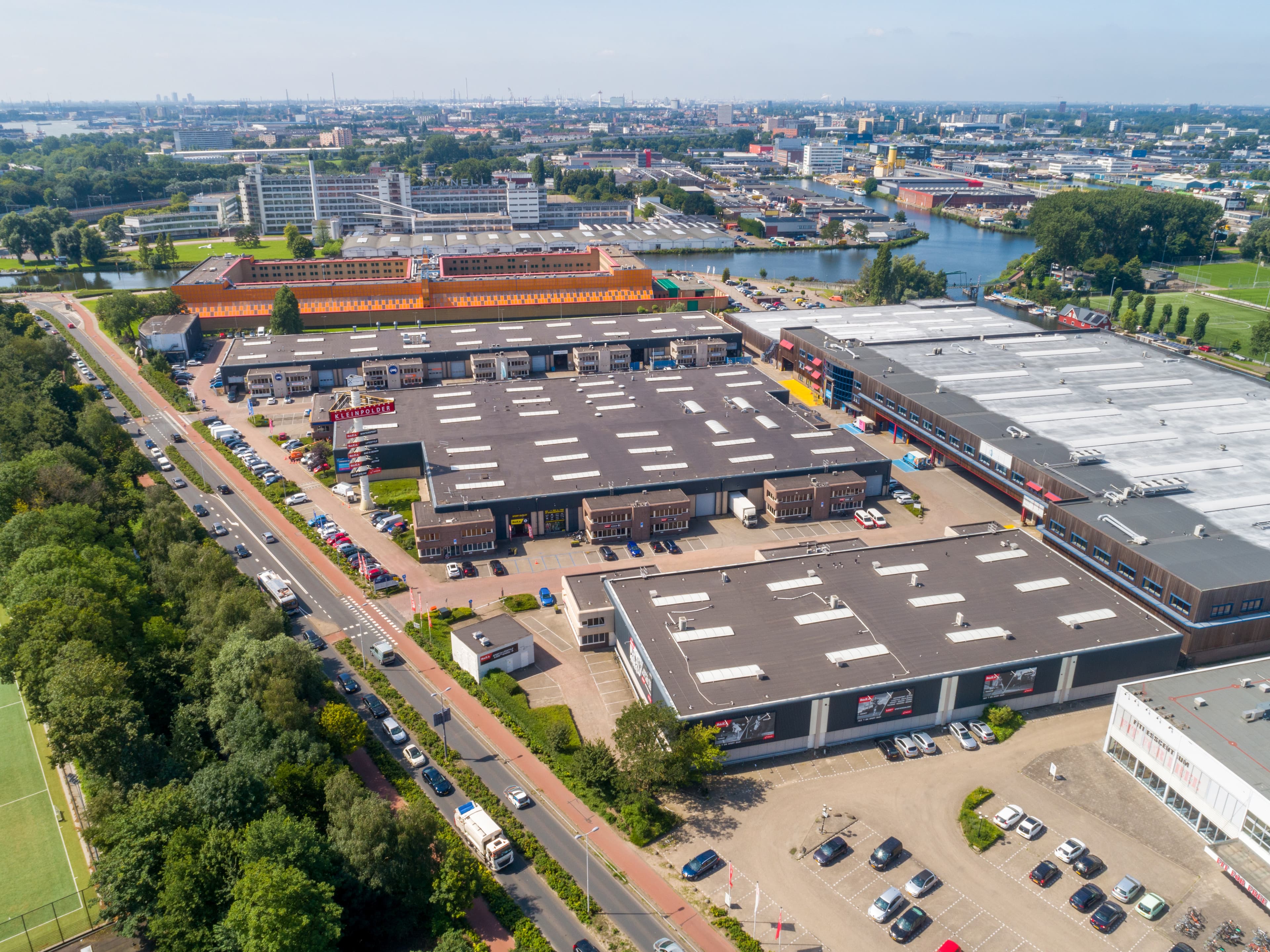 Luftaufnahme eines Industrieparks mit mehreren Lagerhallen, Parkplätzen und Straßen. Im Hintergrund sind städtische Gebäude und ein Fluss unter einem klaren Himmel sichtbar.