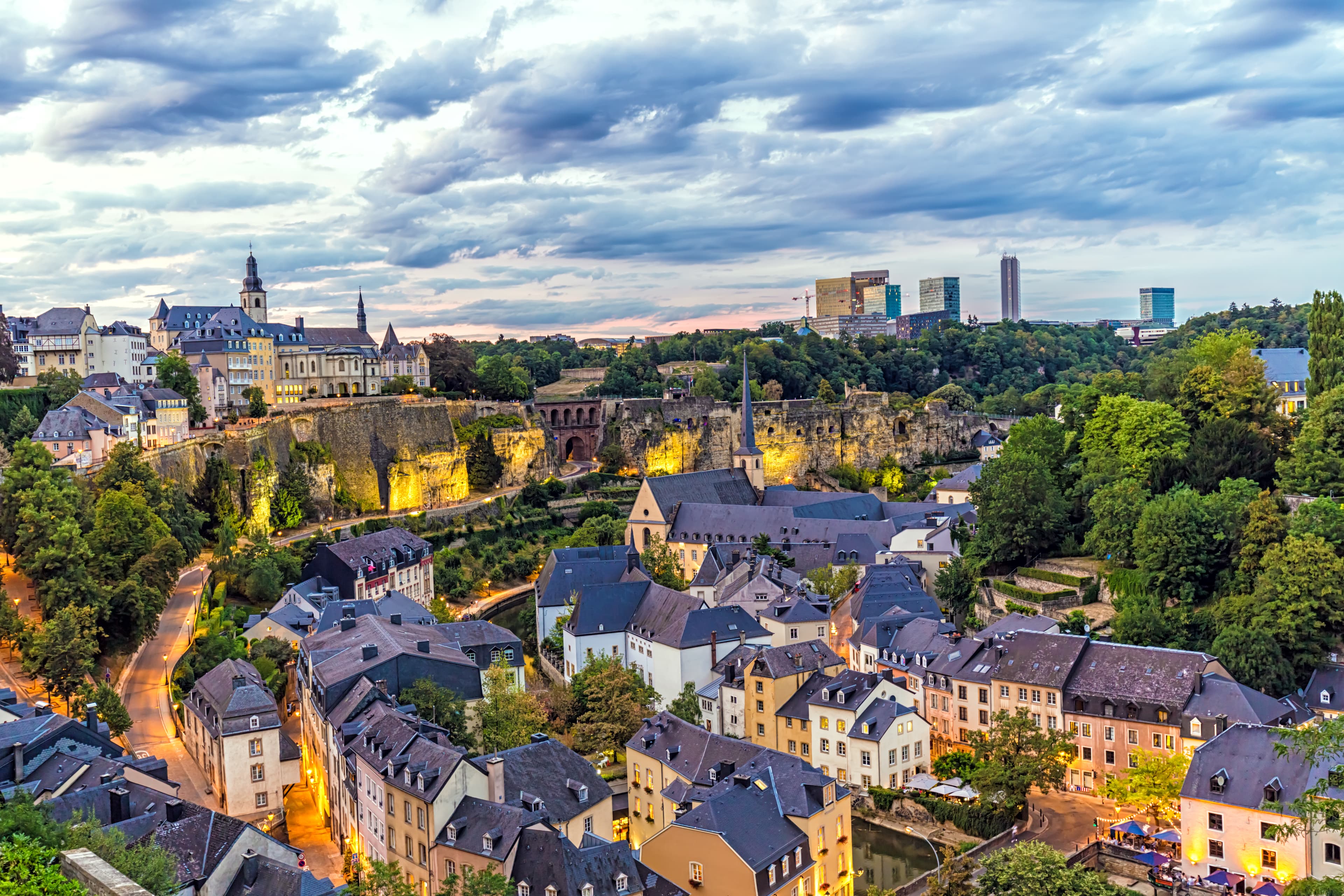 Flygfoto över Luxemburg i skymningen, med historiska byggnader, grönområden och moderna skyskrapor under en molnig himmel.