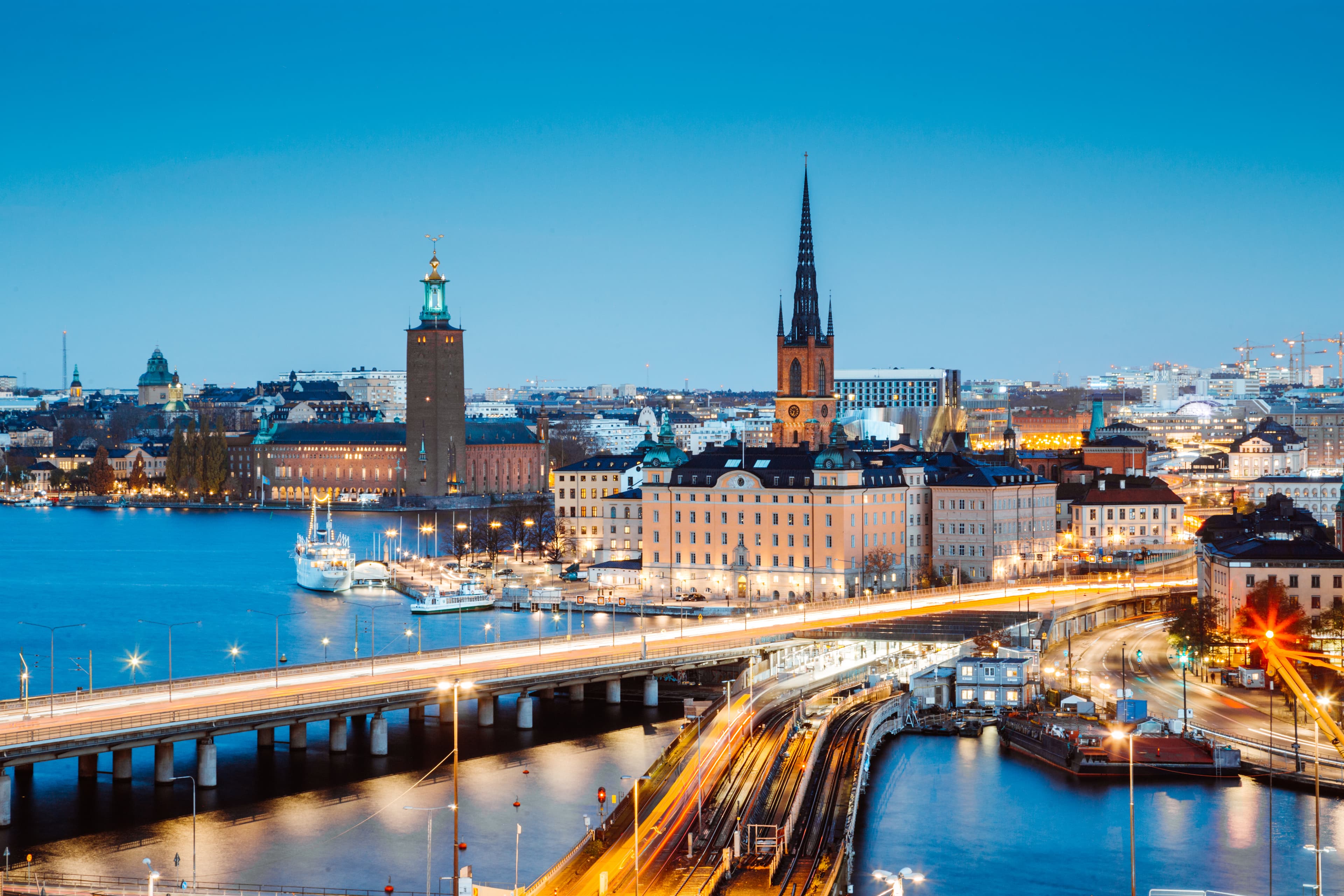 En vy över Stockholm i skymningen, med en upplyst stadsbild med en framstående kyrkspira och byggnader vid vattnet, och suddig trafik på bron i förgrunden.