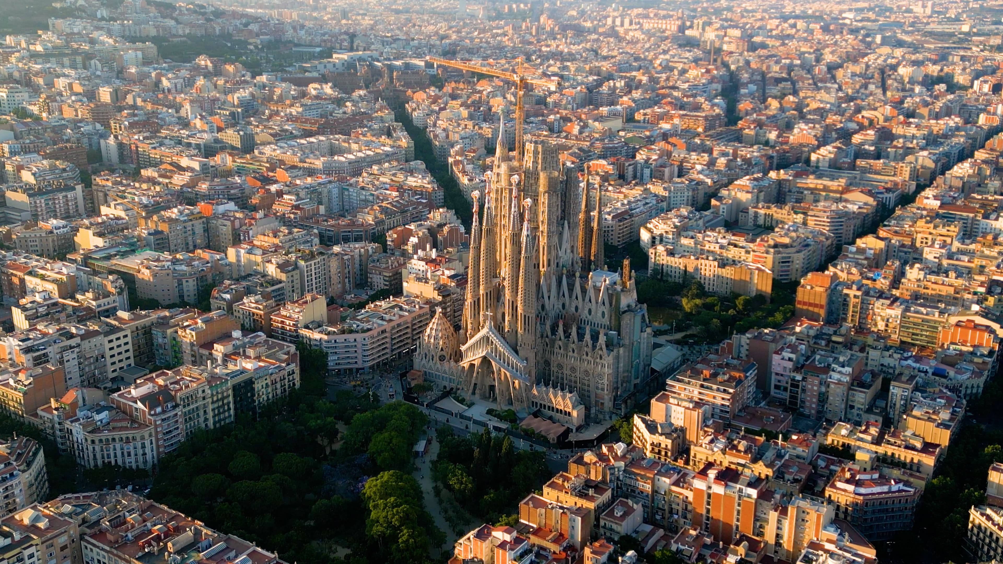 Flygfoto över Sagrada Família i Barcelona, omgiven av stadsgator och byggnader, med en kran synlig ovanpå strukturen.
