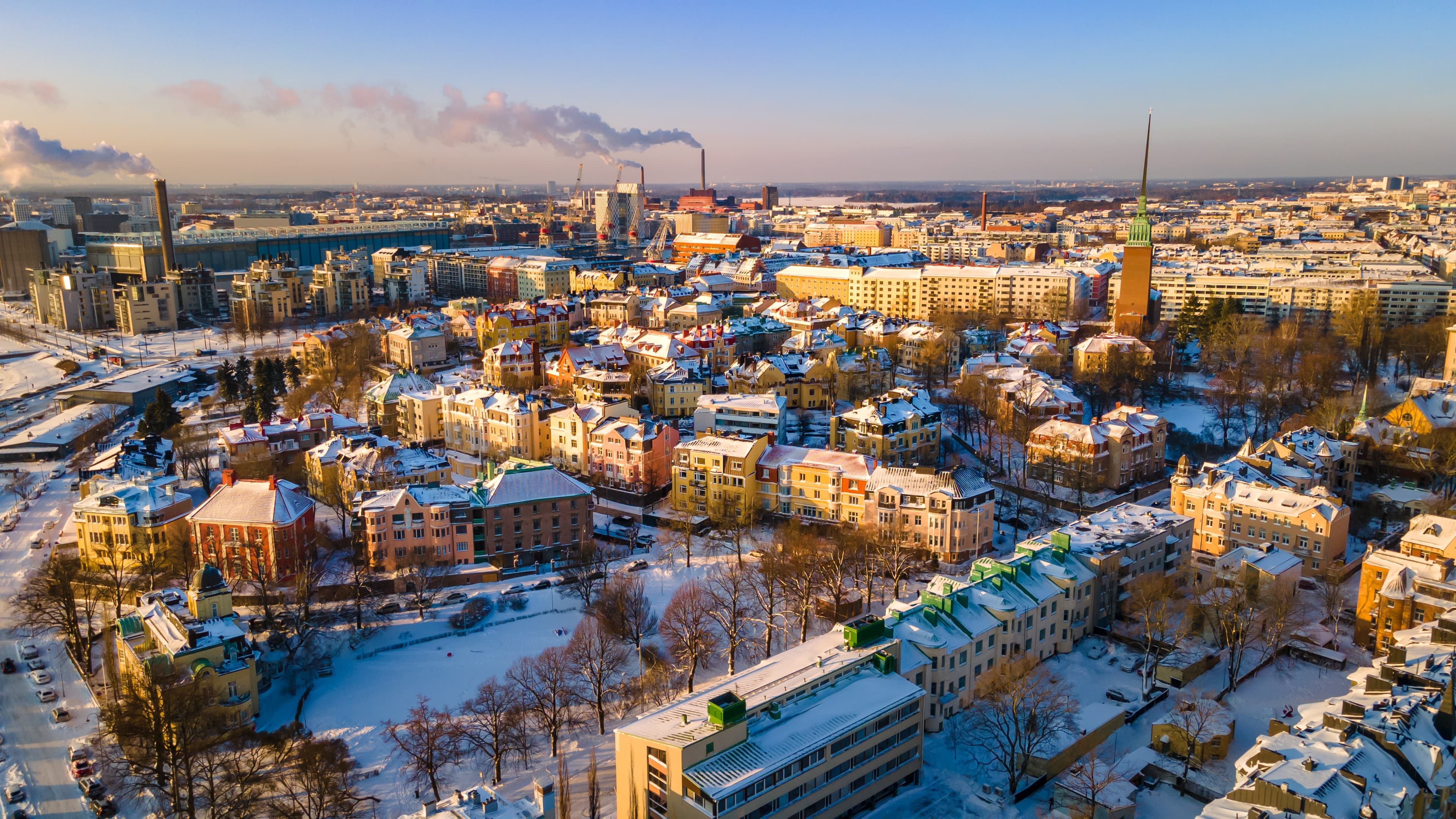 Flygfoto över en snöig stadsbild med färgglada byggnader, kala träd och industriella skorstenar under en klar himmel.