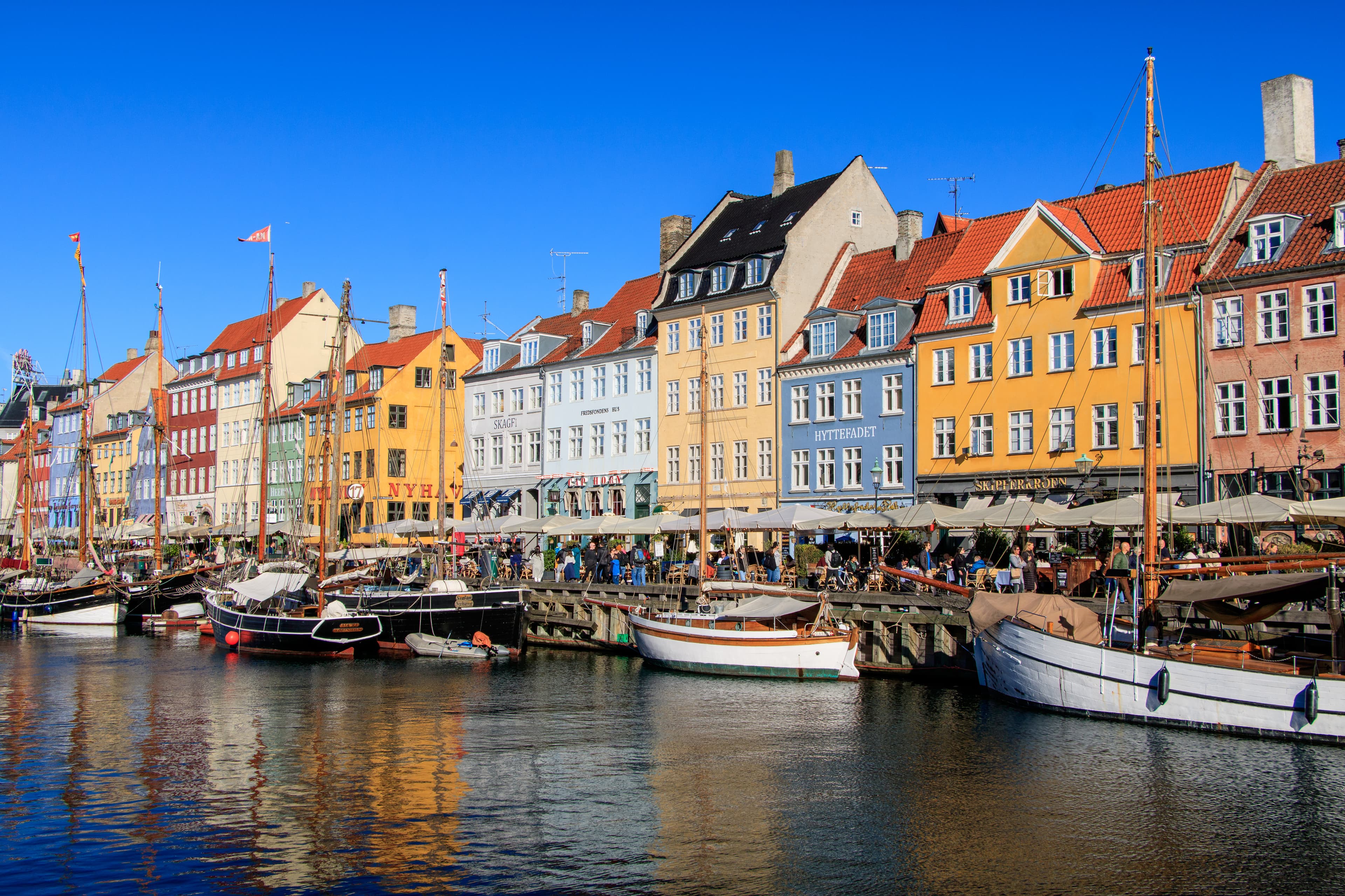 Färgglada byggnader kantar en strand med förtöjda båtar under en klarblå himmel. Scenen speglar sig i det lugna vattnet, vilket skapar en levande och pittoresk utsikt.