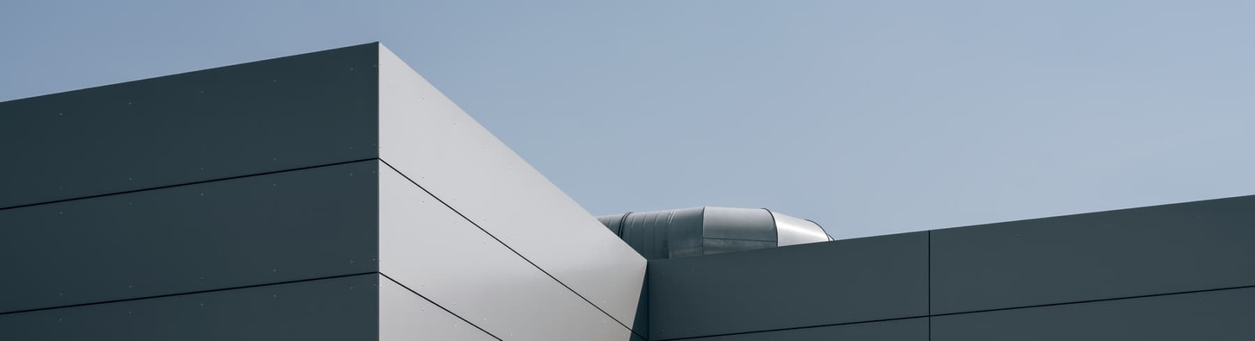 Edificio moderno con paneles de metal gris y una estructura cilíndrica en el techo contra un cielo azul claro.