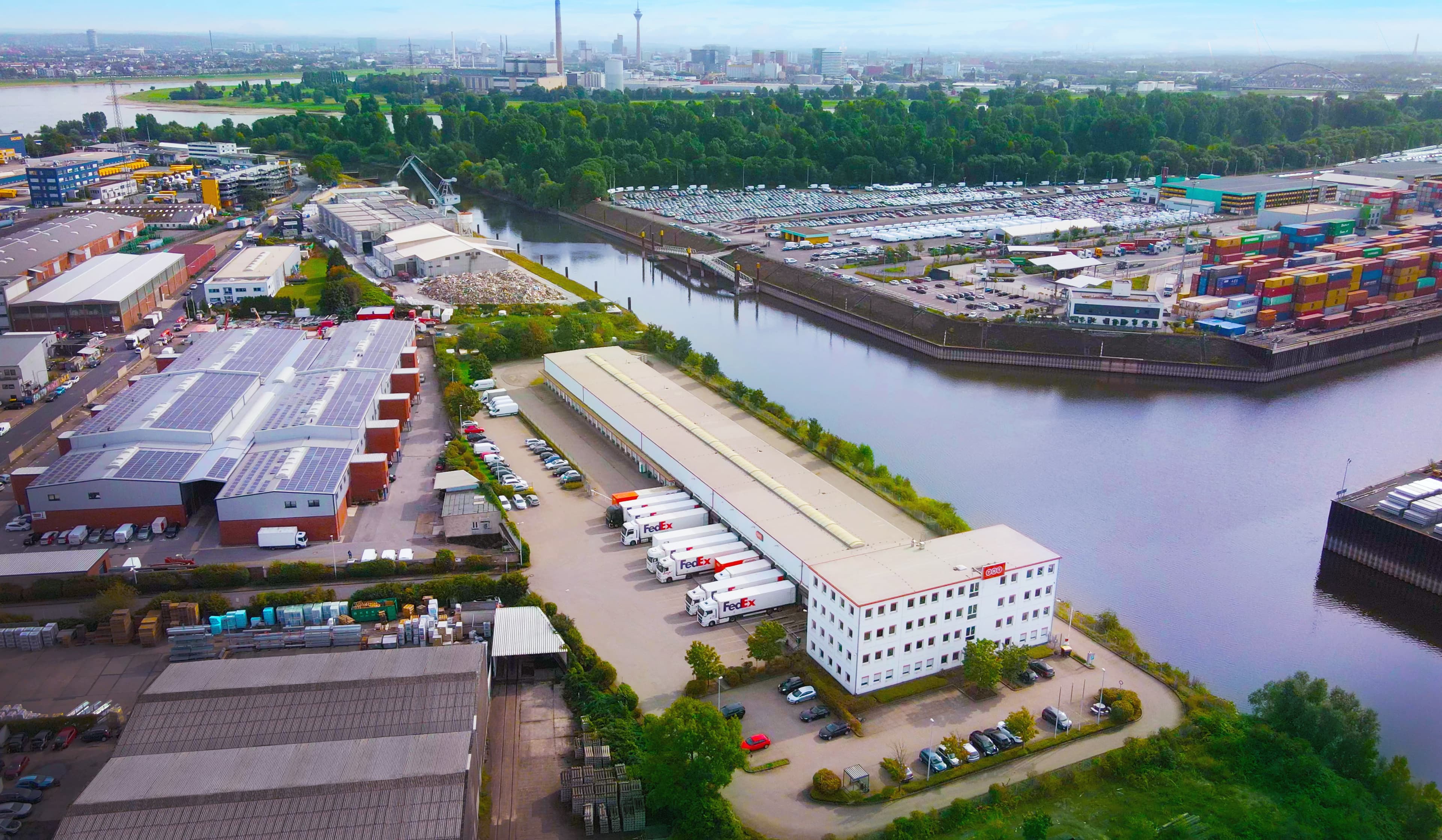 Luftaufnahme eines Industriegebiets neben einem Fluss mit Lagerhallen, einer FedEx-Anlage mit geparkten Lastwagen, Containerlager und Stadtbild im Hintergrund.