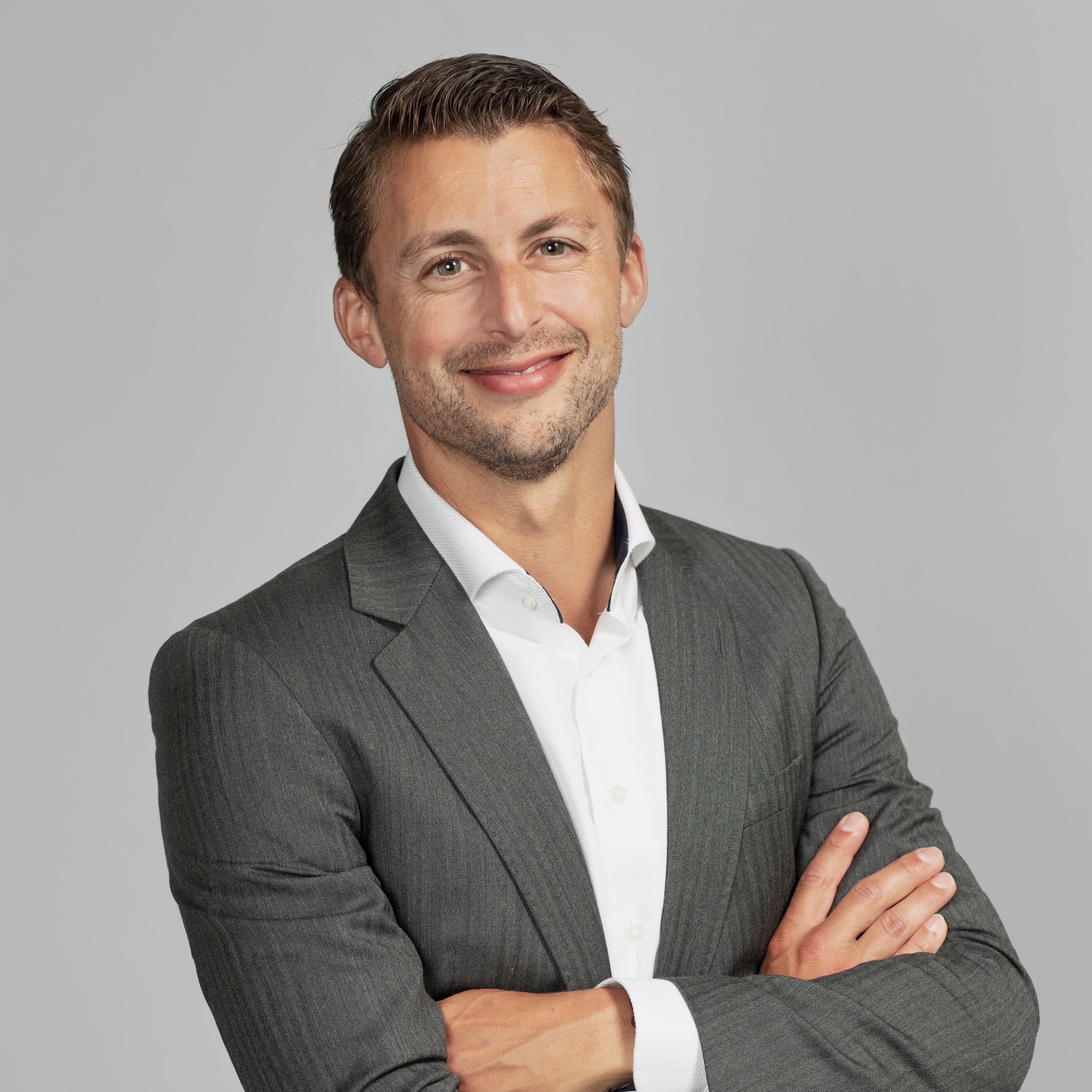 Een man in een grijs pak en een wit overhemd staat met gekruiste armen en lacht tegen een effen achtergrond.