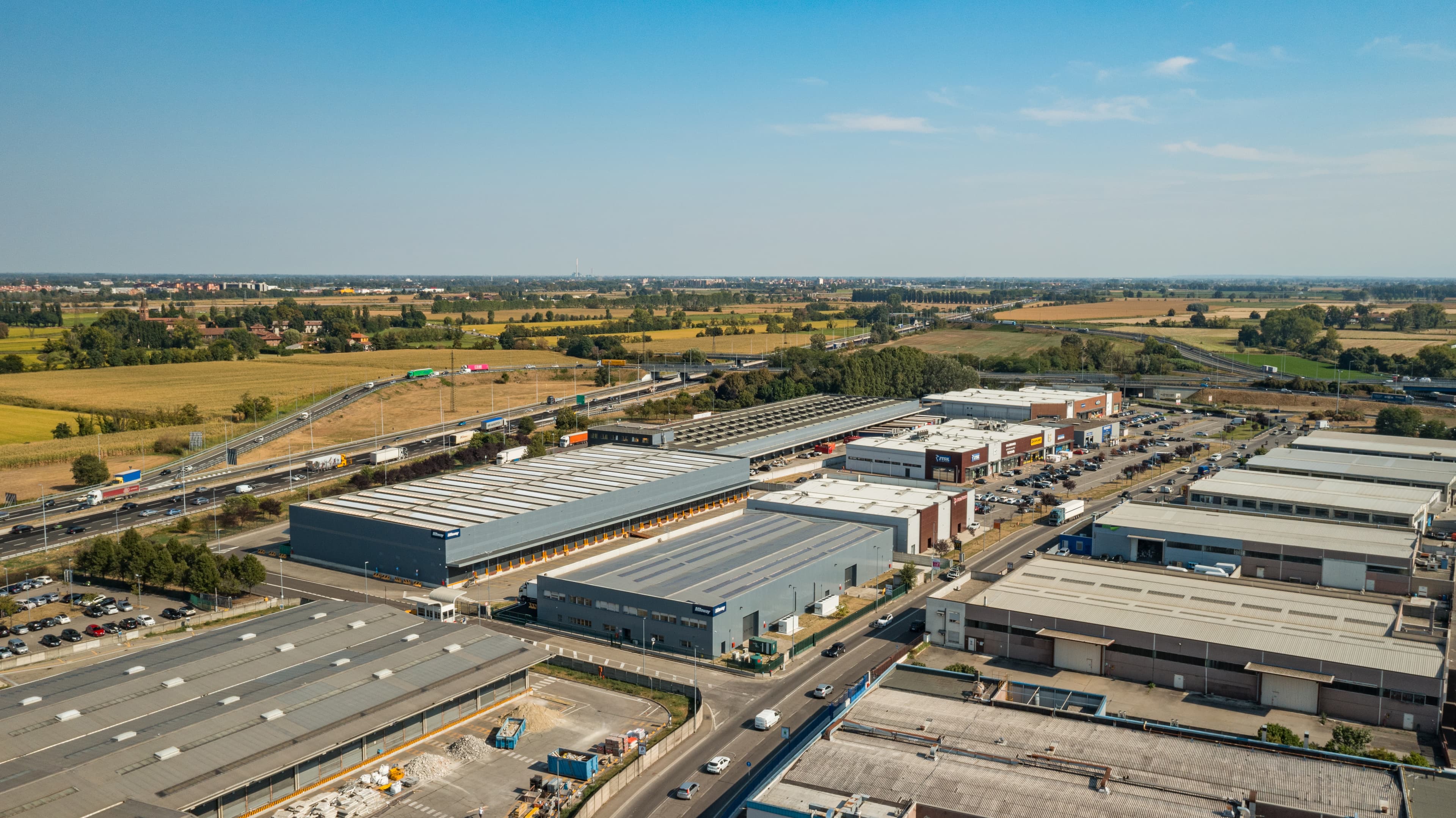 Vista aerea di una zona industriale con numerosi magazzini e fabbriche circondati da strade e campi sotto un cielo azzurro e limpido.