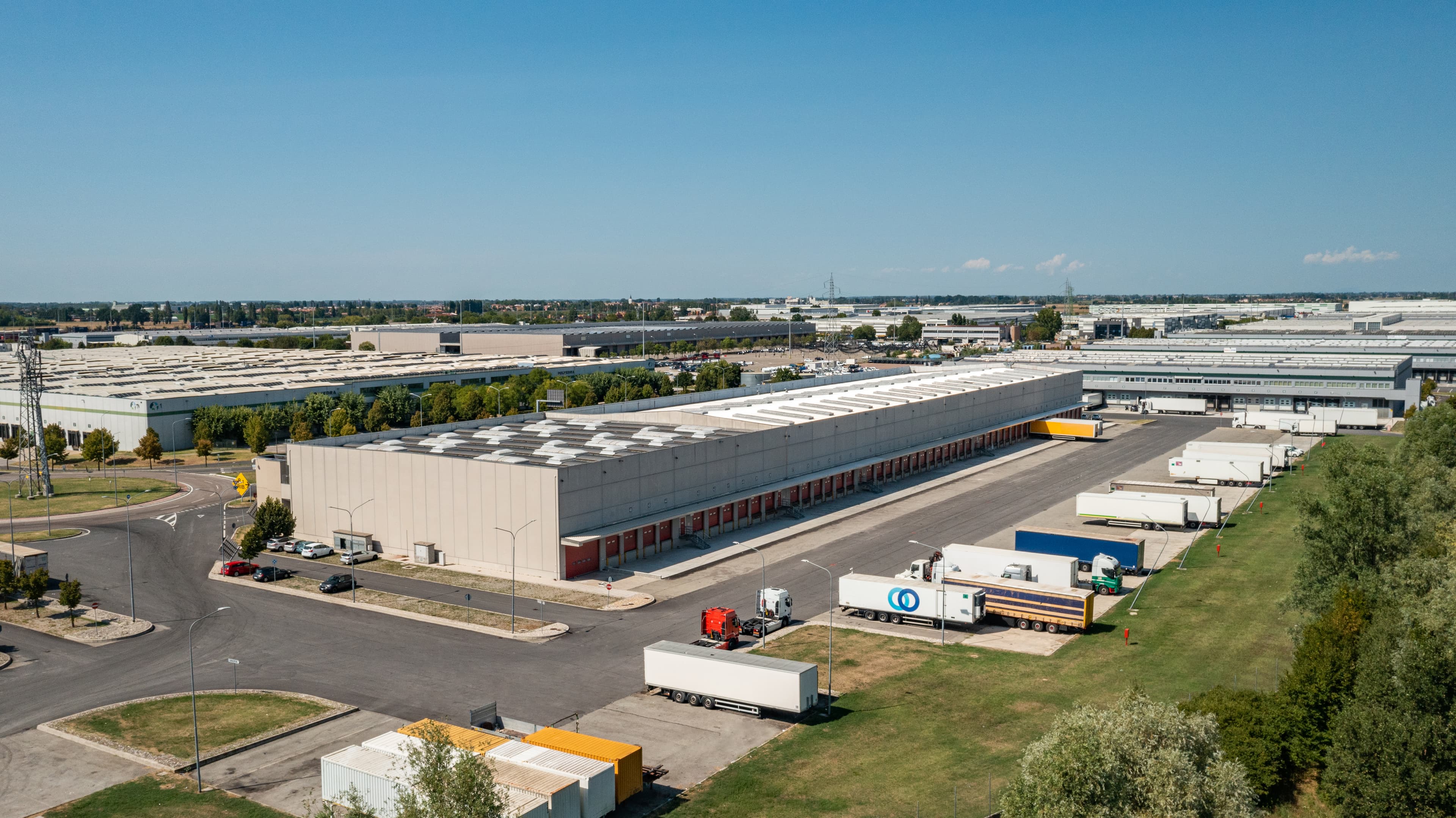 Vista aerea di un grande complesso industriale con numerosi magazzini circondati da camion e verde sotto un cielo azzurro e limpido.
