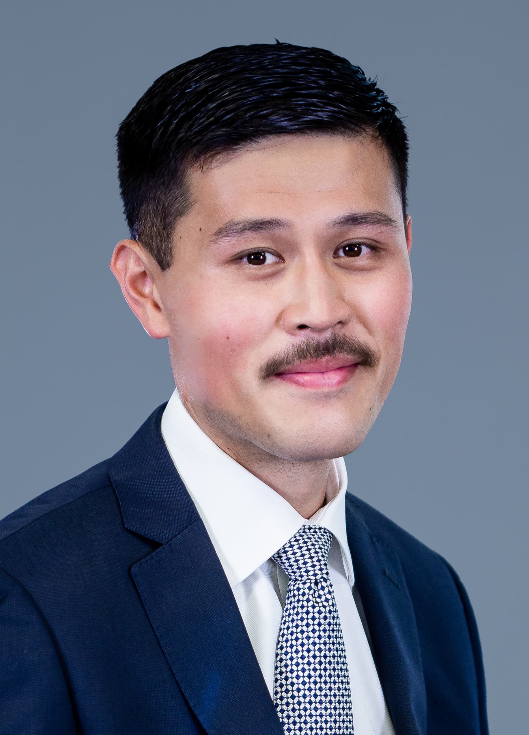 A man in a dark suit, white shirt, and patterned tie poses for a professional portrait against a plain grey background.