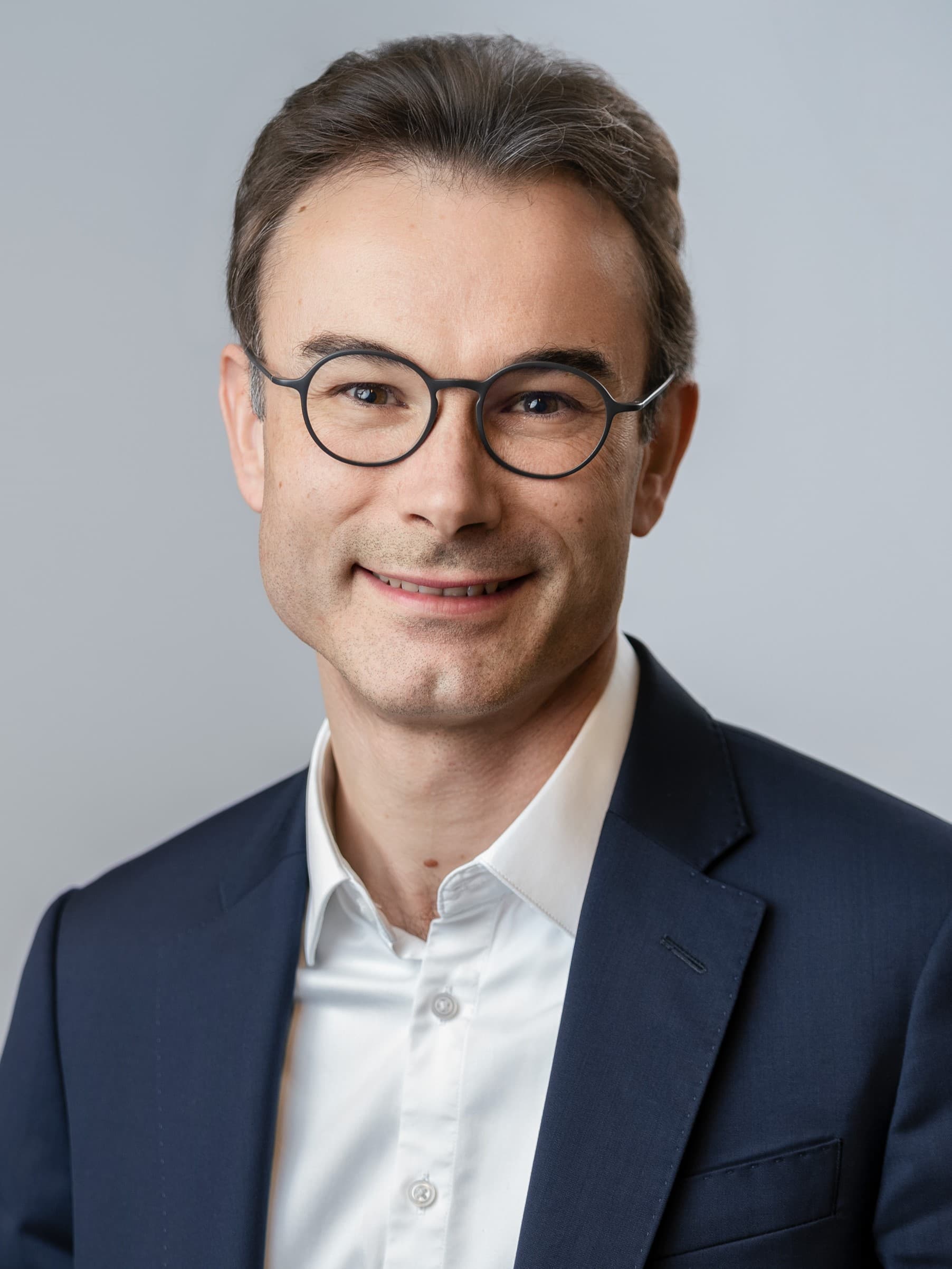 A man with short brown hair, wearing glasses, a white shirt, and a dark suit jacket, smiling at the camera against a plain background.