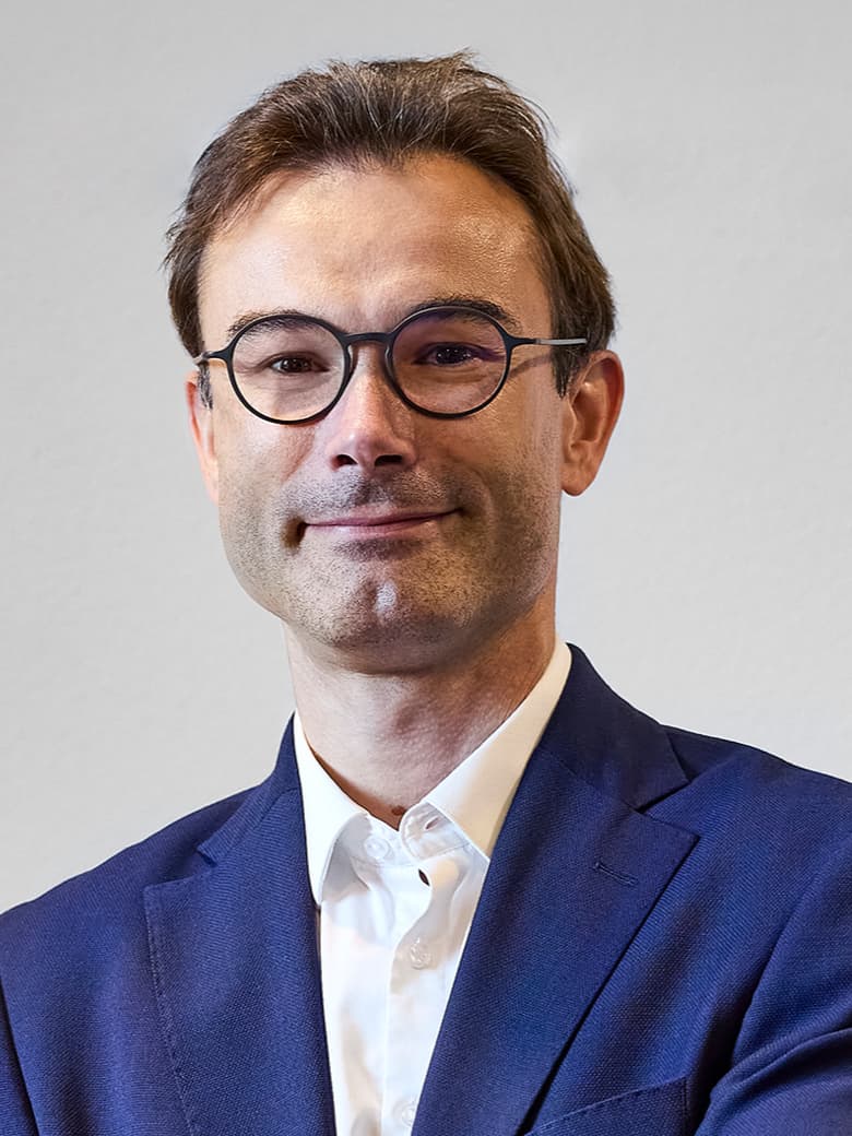 A man with short hair is wearing round glasses, a blue suit, and a white shirt. He is facing the camera with a slight smile against a plain background.