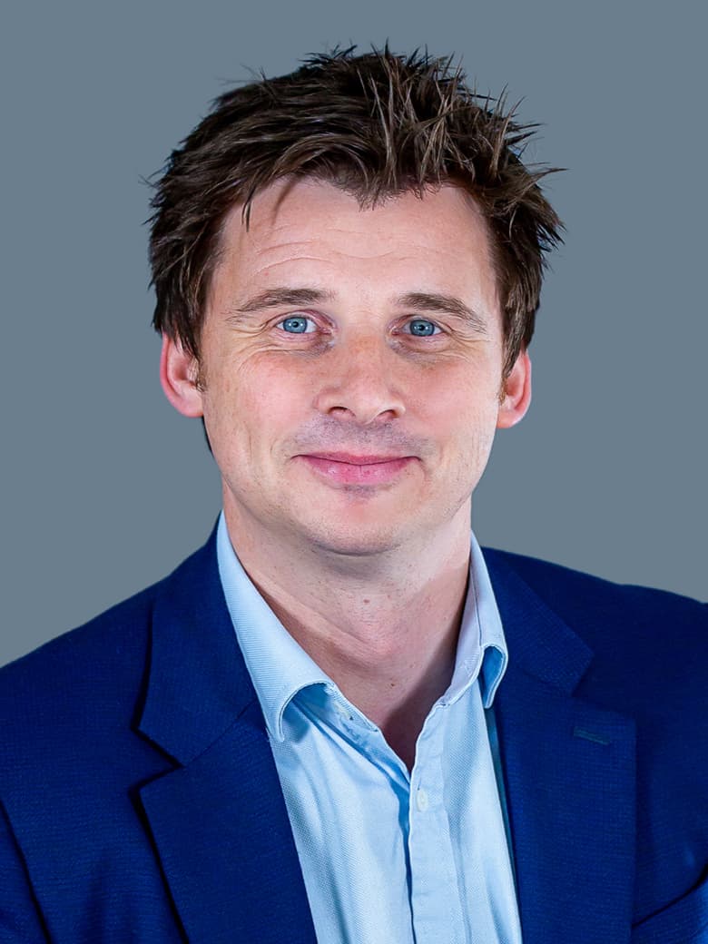 A person with short brown hair wearing a blue suit jacket and light blue shirt, smiling slightly, against a plain gray background.