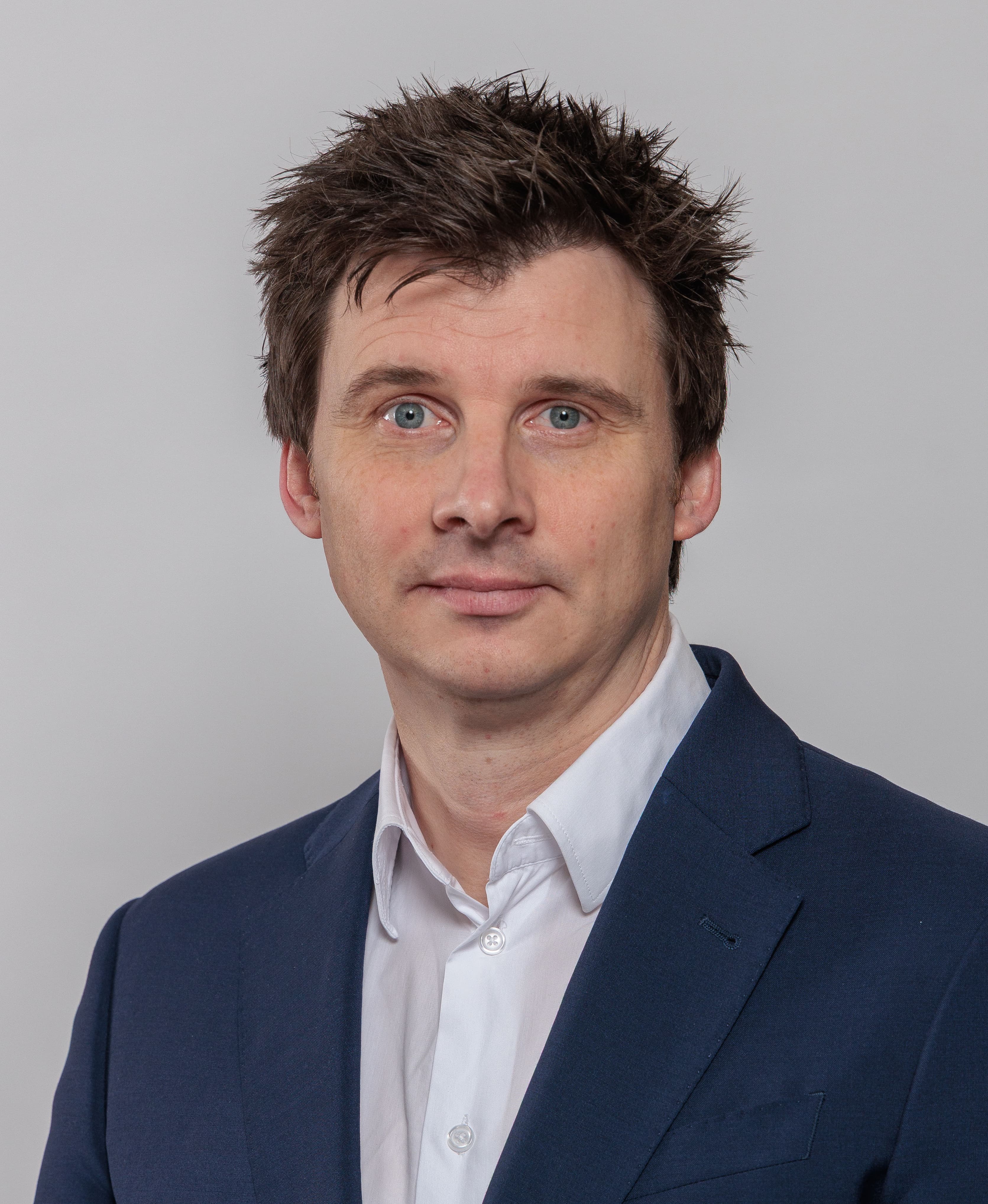 A man with short brown hair, wearing a navy blue suit and white shirt, stands against a plain light grey background.