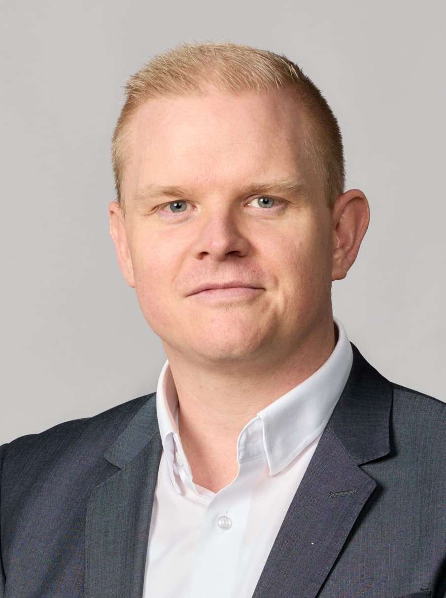 A person wearing a dark suit jacket and white dress shirt posing against a light grey background.