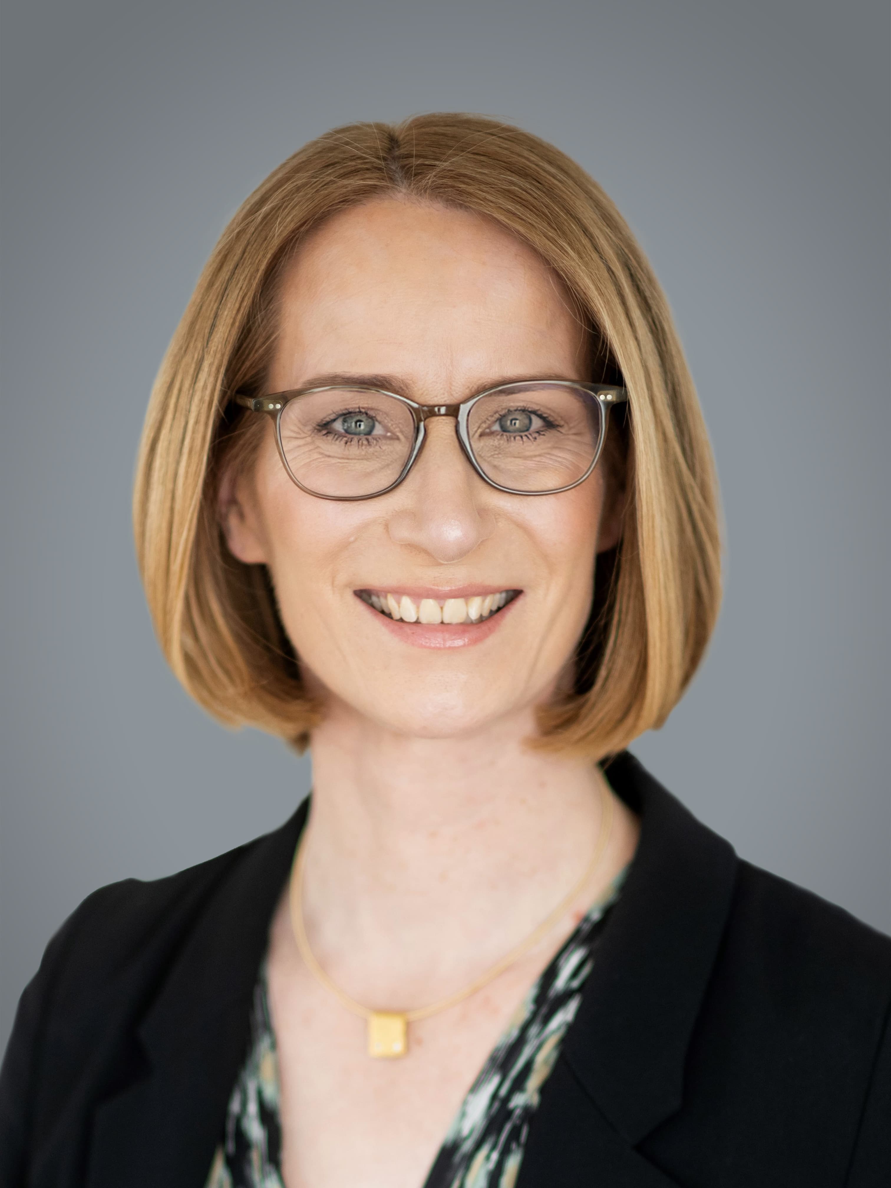 A person with short, light brown hair and glasses smiles, wearing a black blazer and a patterned top against a grey background.