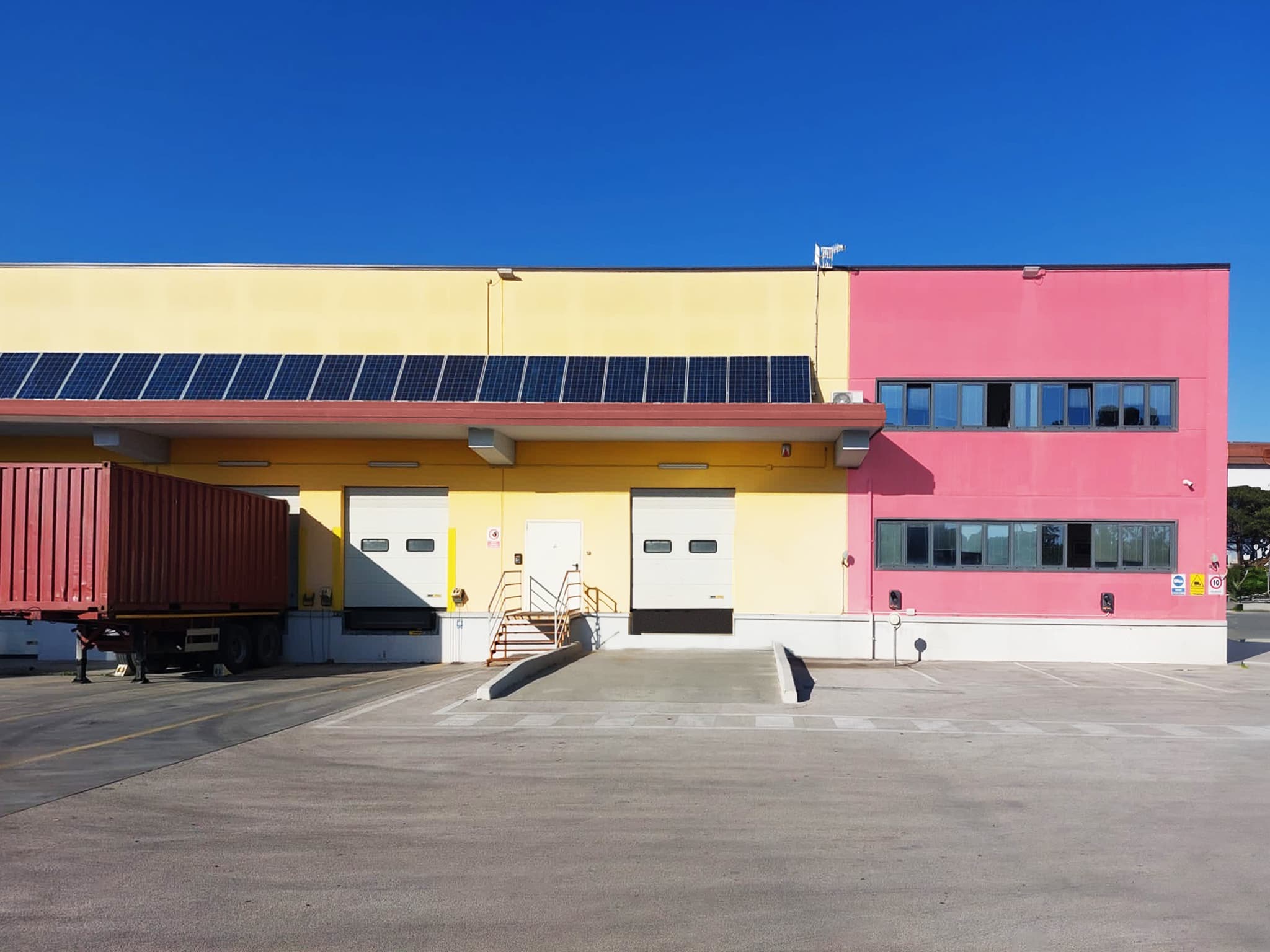 L'esterno colorato di un magazzino presenta pannelli solari sul tetto. Un container rosso è attaccato a un rimorchio di fronte a due grandi baie di carico.
