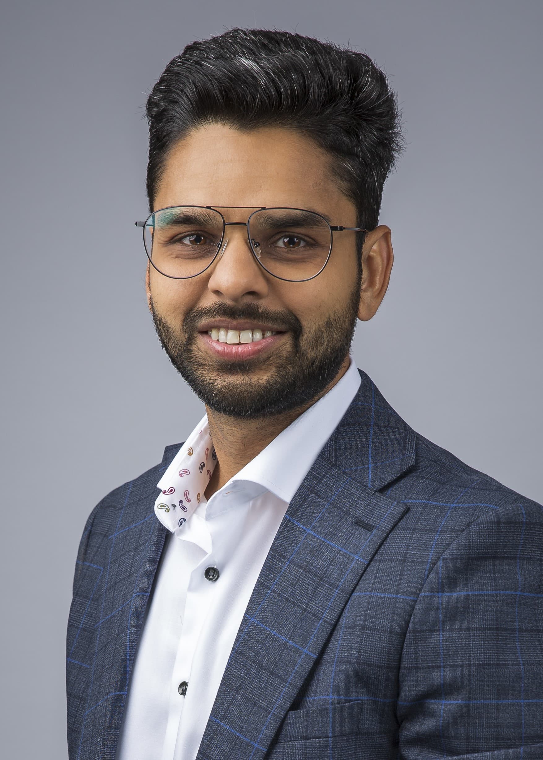 A person with glasses and a beard smiles slightly, wearing a checked suit jacket and white shirt against a gray background.