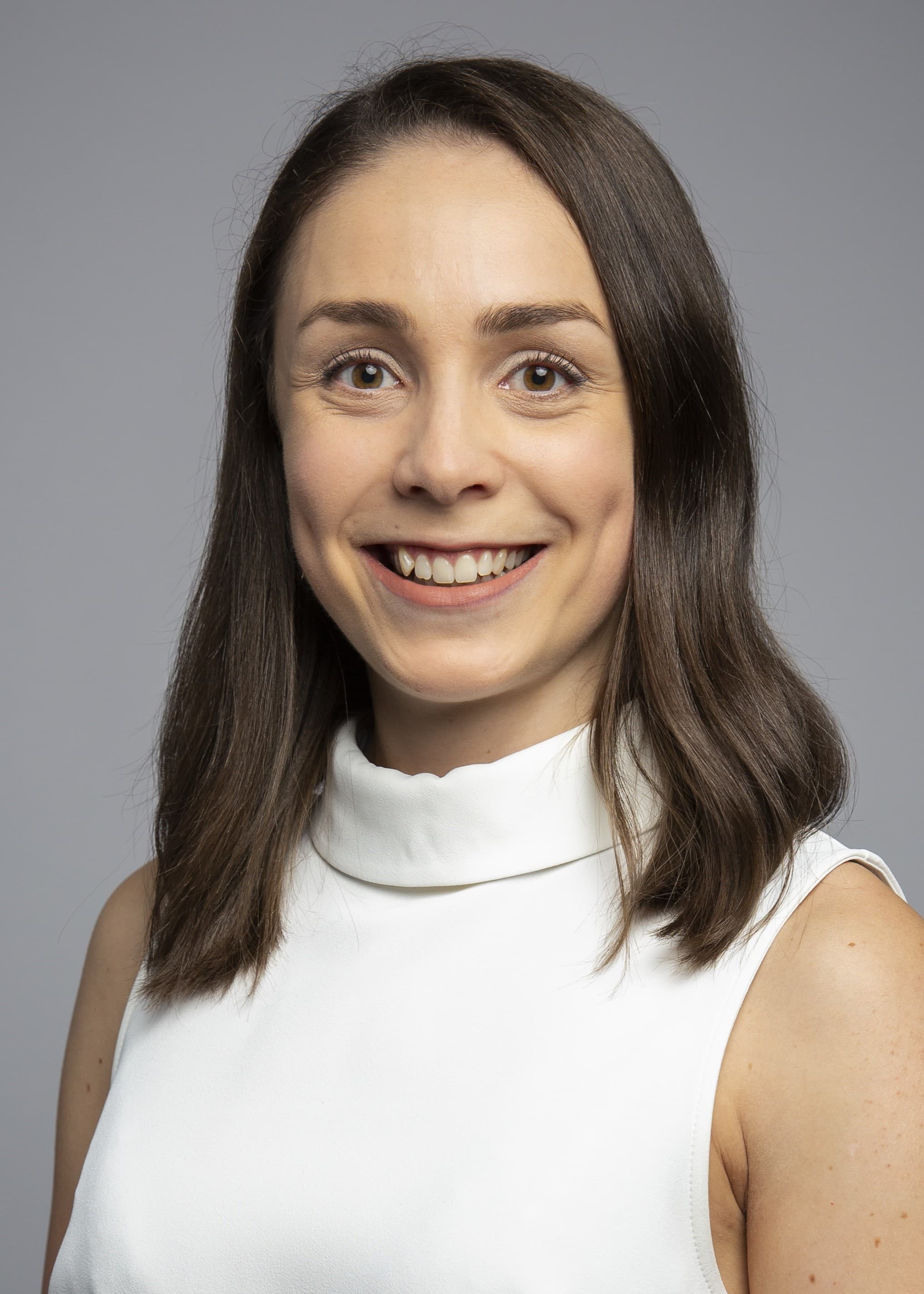 A person with shoulder-length brown hair wearing a sleeveless white top smiles at the camera against a gray background.