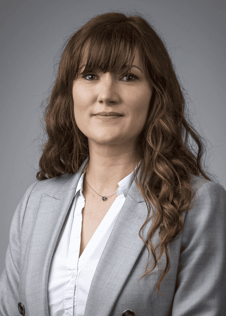 A woman with long brown hair wears a light gray blazer and white shirt, posing against a neutral gray background.