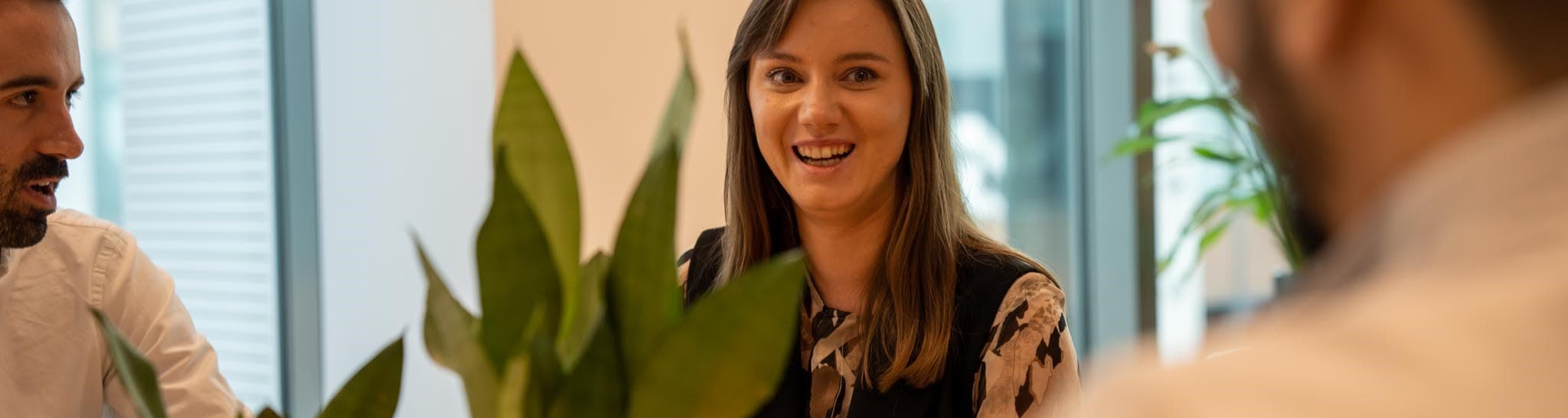 Tre persone sono sedute a un tavolo con una pianta in primo piano, e stanno conversando. L'attenzione è rivolta alla persona al centro che sorride.