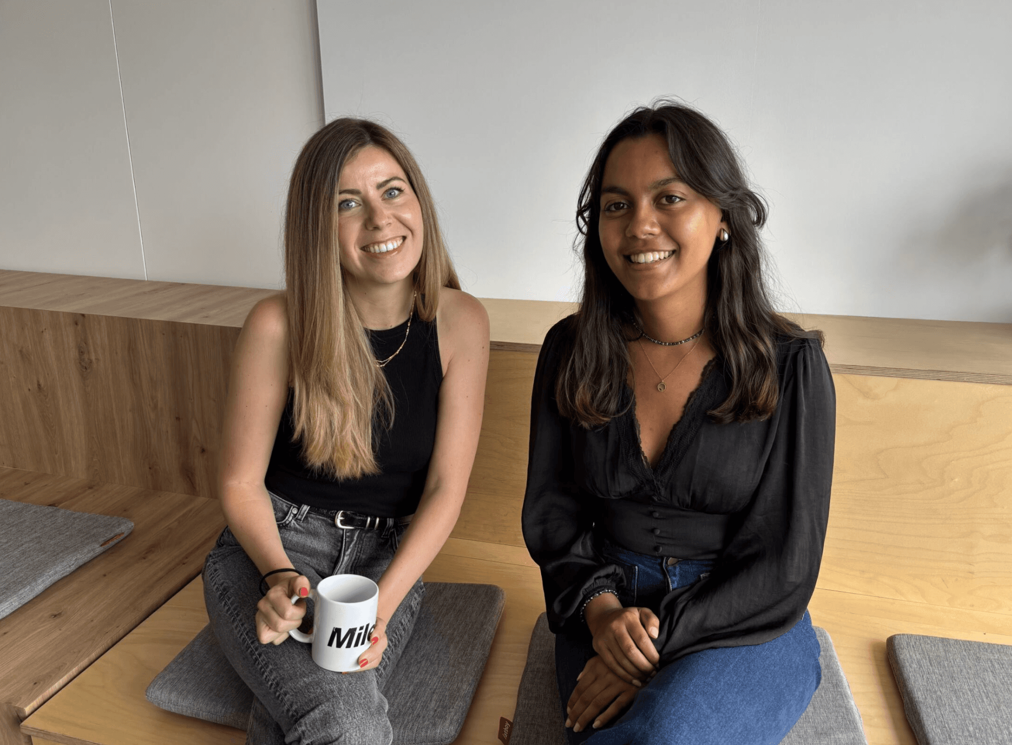 Two women sitting on wooden benches indoors, smiling at the camera. One holds a white mug labeled Milk. Both wear black tops and have long hair.