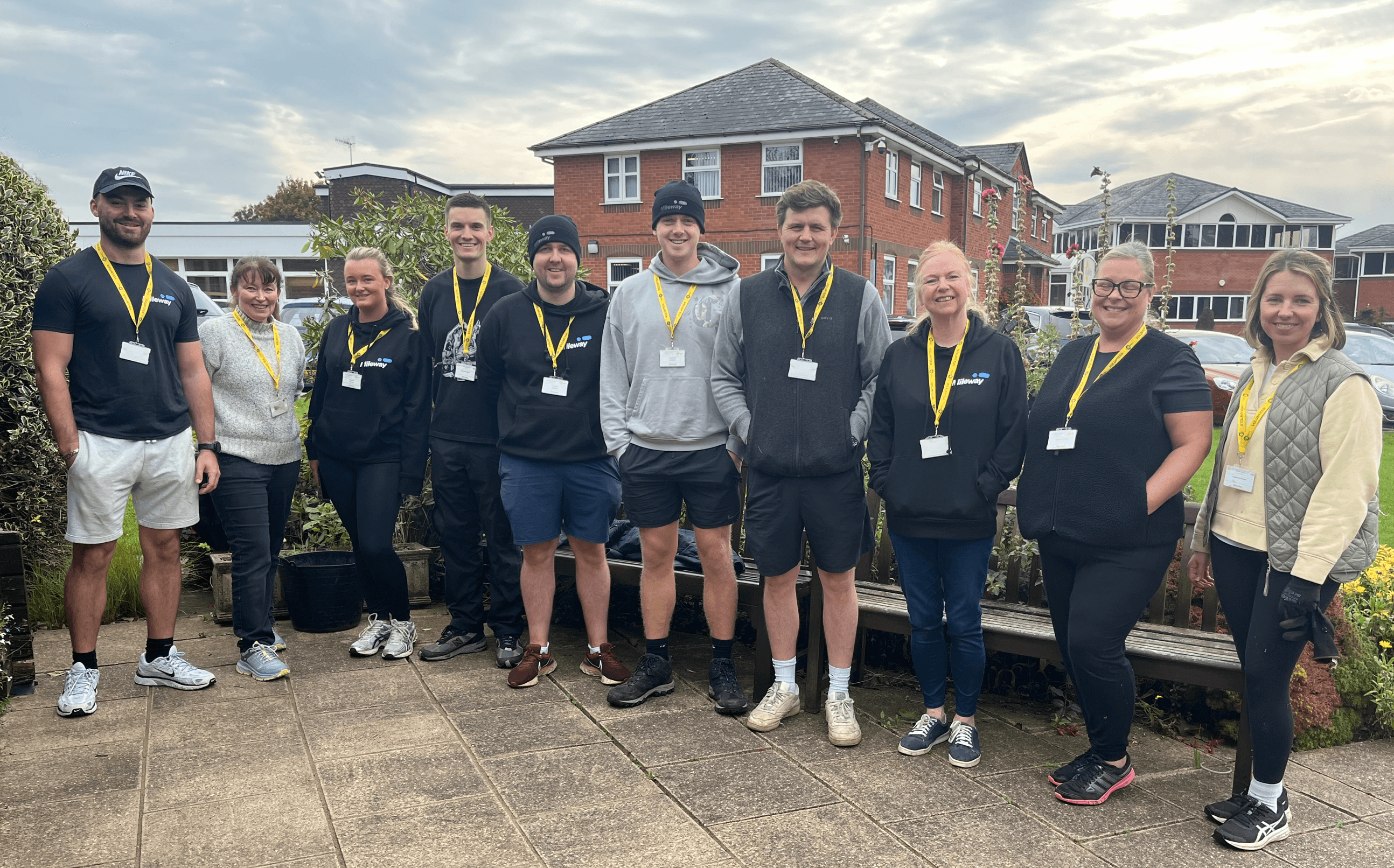 Ten people wearing lanyards stand in a row outdoors in front of a building, posing for a group photo on a cloudy day.
