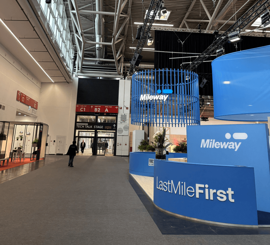 A modern exhibition hall featuring blue Mileway stands, Last Mile First signage, and a Tech Talk Stage entrance in the background.