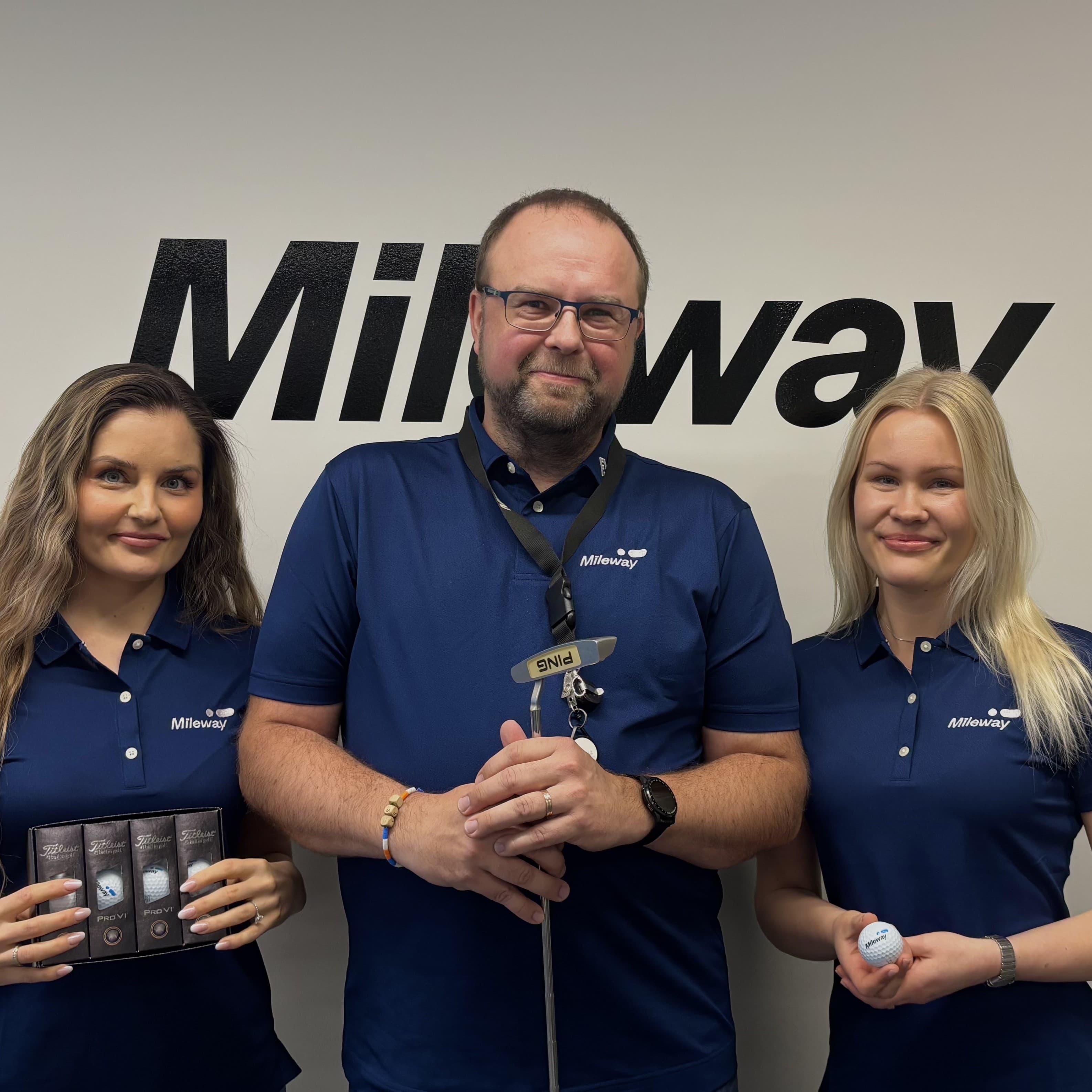 Three people in matching blue Mileway shirts stand in front of a Mileway logo; the man in the middle holds a golf club, while the women hold chocolate and a golf ball.