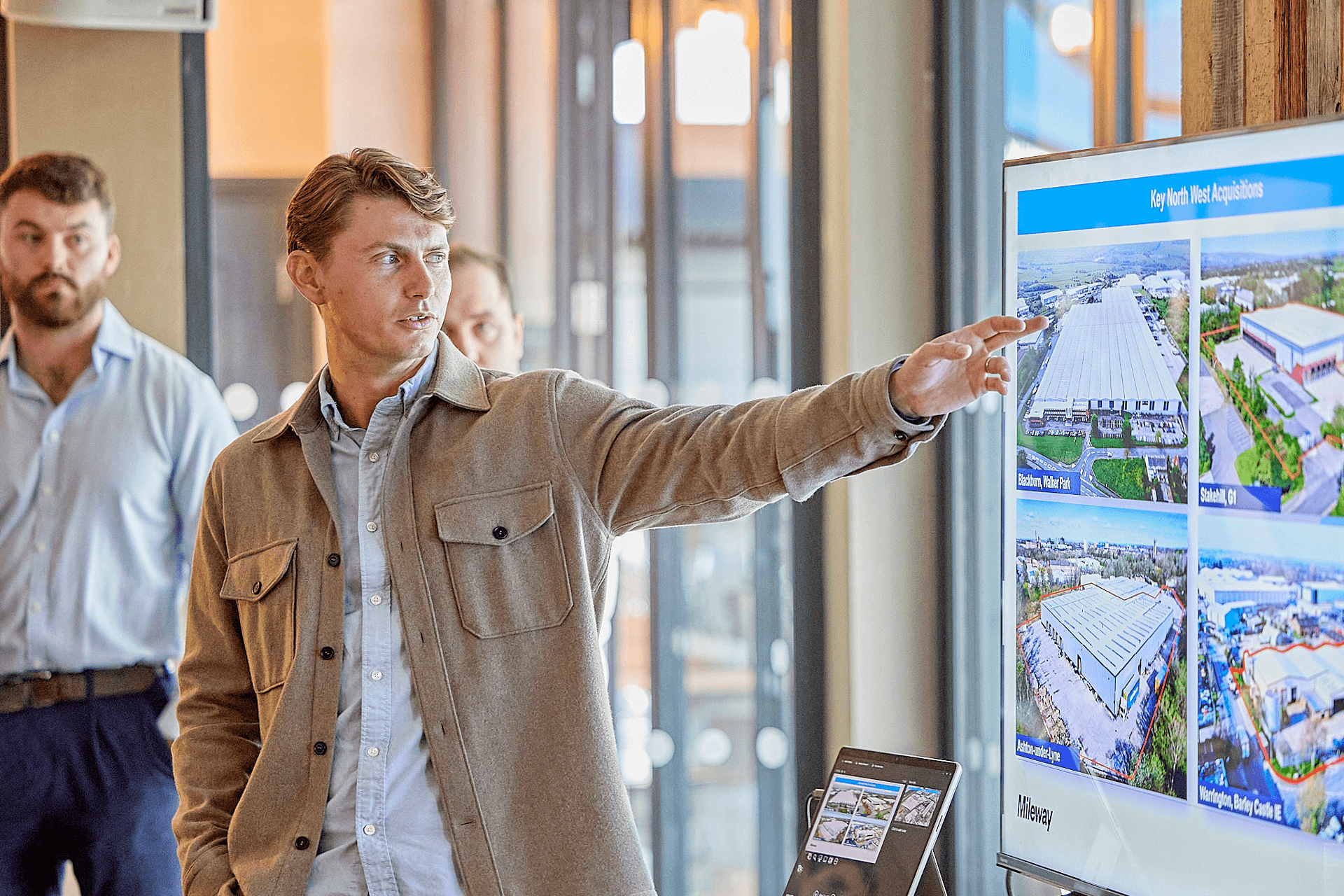 A man in a beige jacket points to a large screen displaying images and text during a business presentation, with two other men in the background.