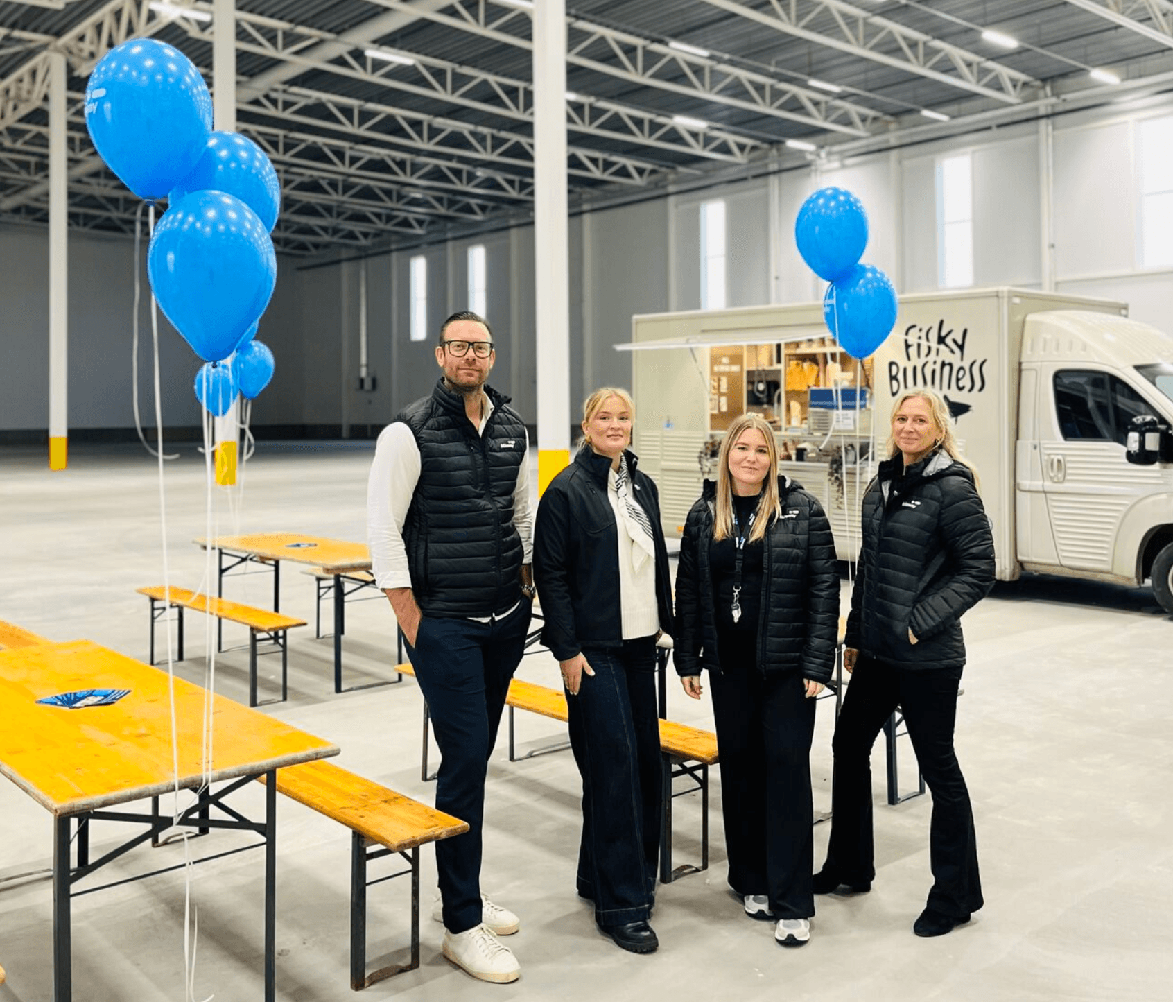 Four people in black waistcoats stand indoors beside wooden tables with blue balloons; a food van labelled “Fisky Business” is in the background.