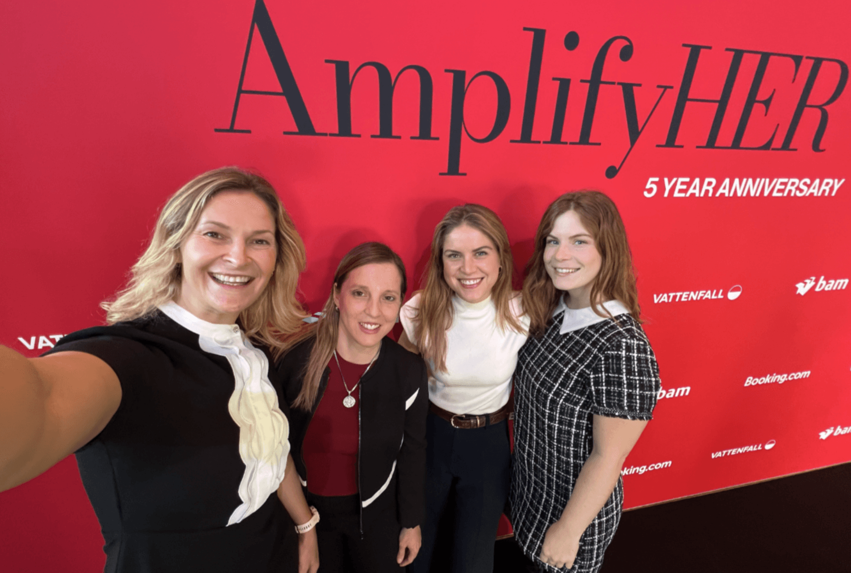 Four women stand together, smiling for a group selfie in front of a red AmplifyHER 5 Year Anniversary event backdrop.