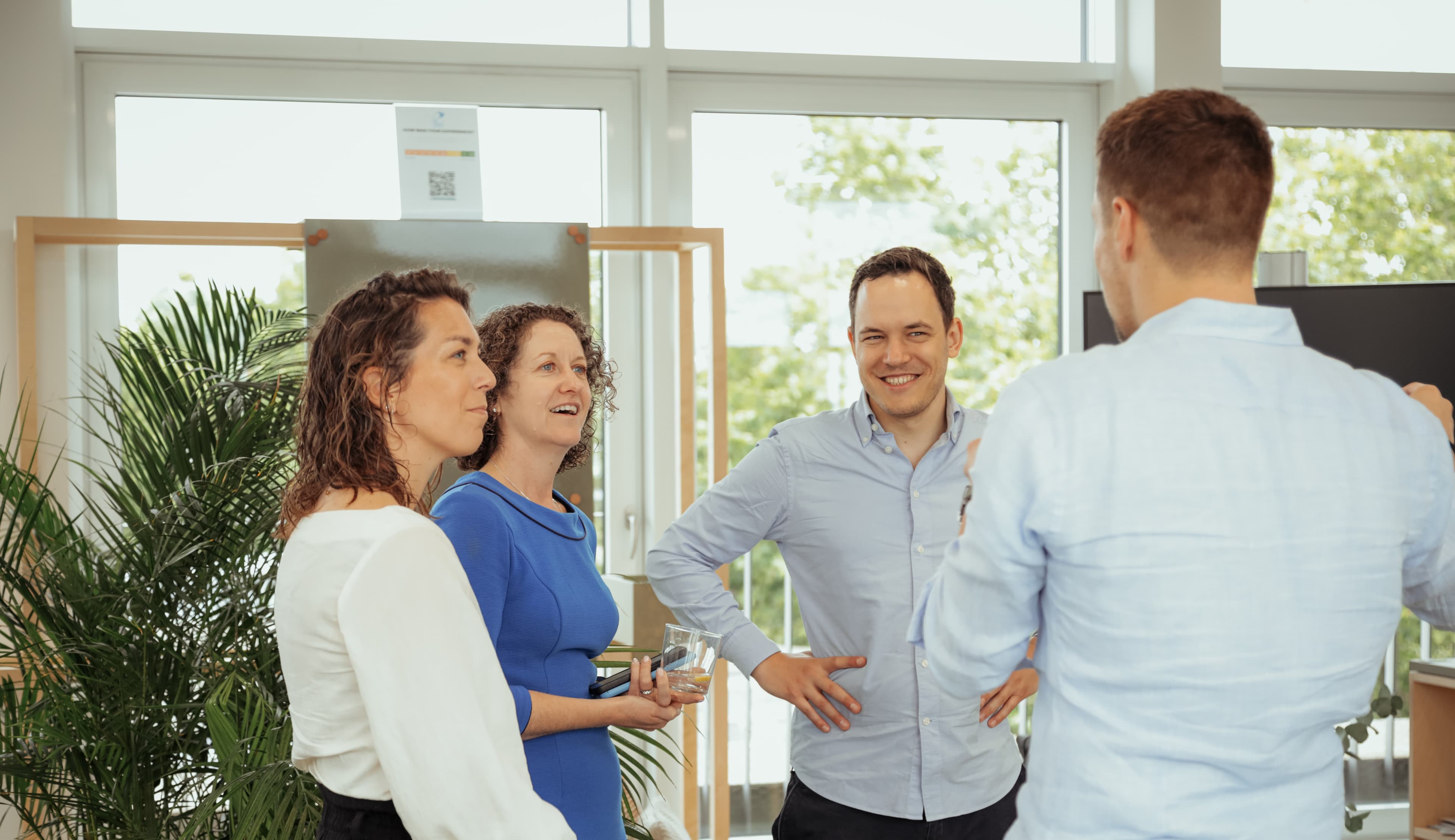 Vier Personen stehen in einem Innenraum zusammen und unterhalten sich. Drei von ihnen sind der Kamera zugewandt, eine hat ihr den Rücken zugewandt. Im Hintergrund sind große Fenster und Grünpflanzen zu sehen.