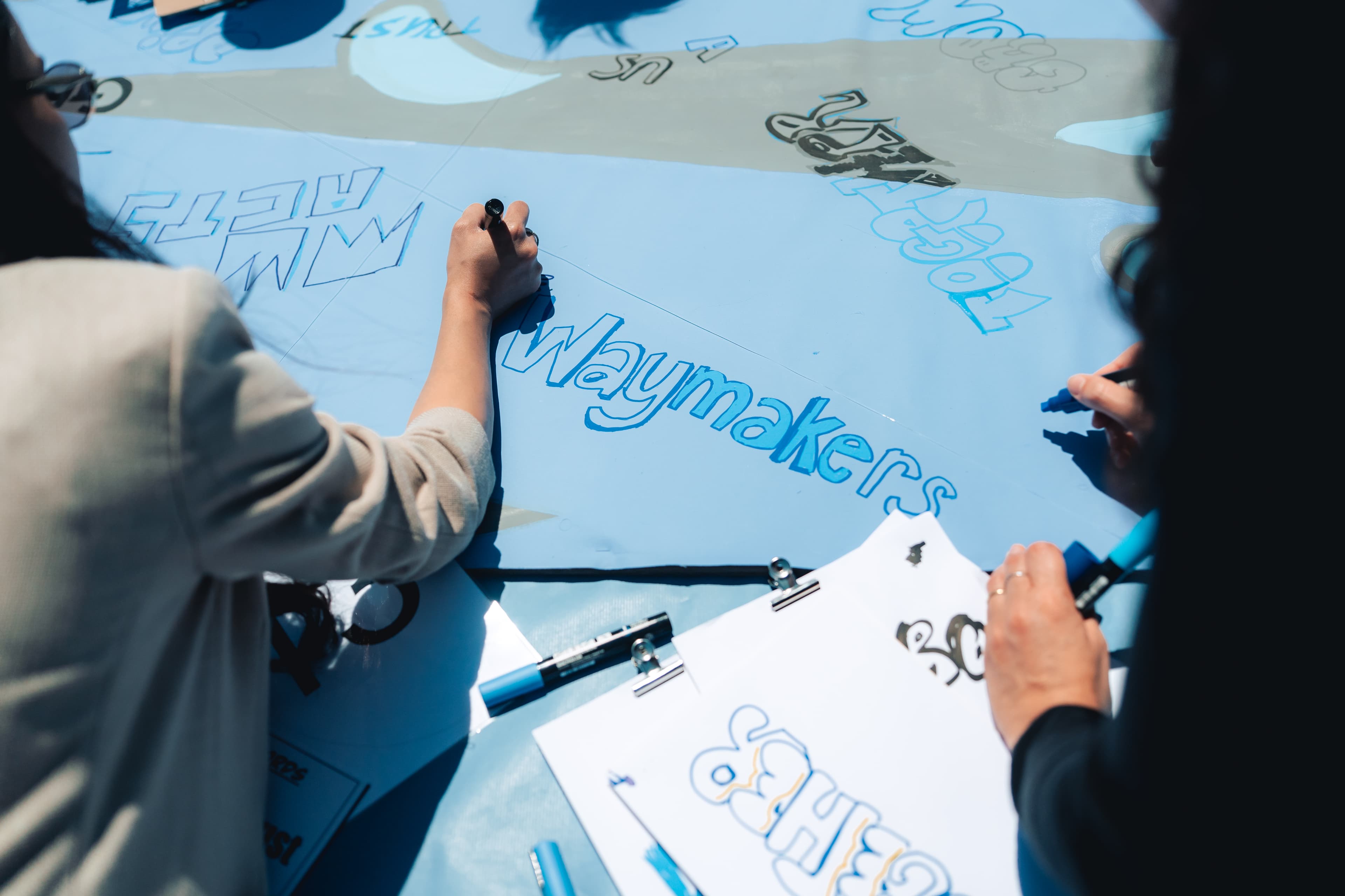 Dos personas utilizan rotuladores para escribir y dibujar palabras estilizadas, entre ellas "waymakers", en un gran papel azul rodeado de portapapeles y papel.