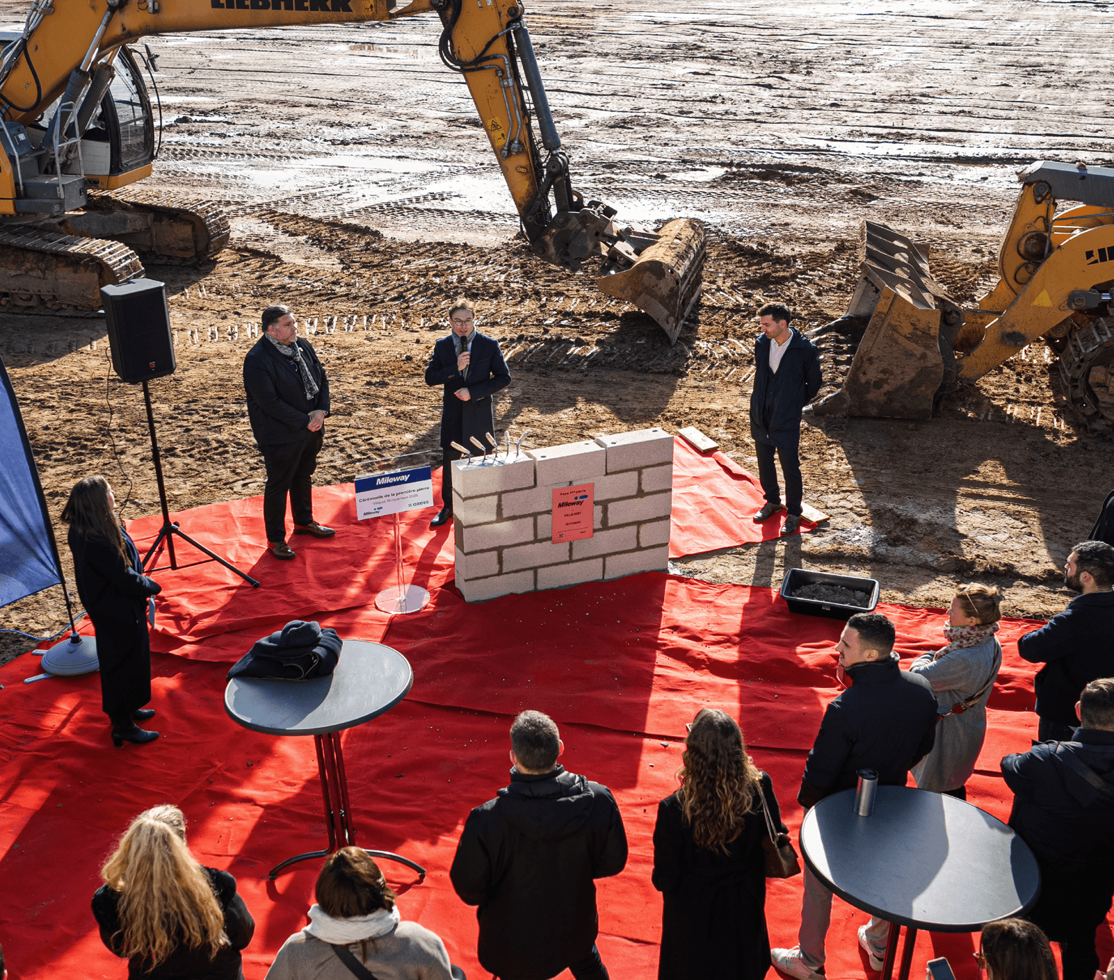 A group of people watch a speaker at a construction site event, with a brick wall display and diggers in the background on a red carpeted area outdoors.