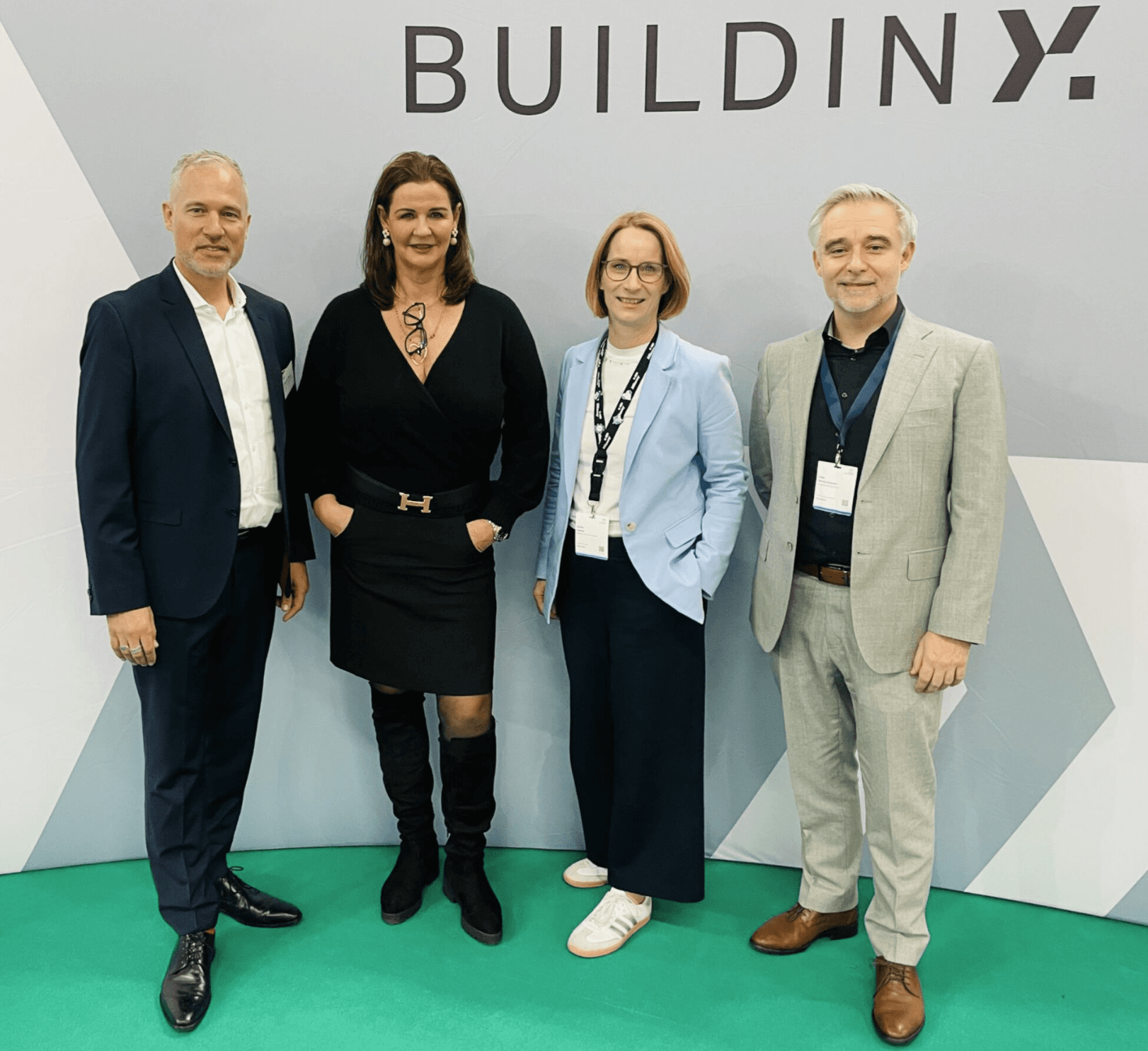 Four professionally dressed adults stand in front of a wall with the text BUILDINX, posing for a group photo at an event.