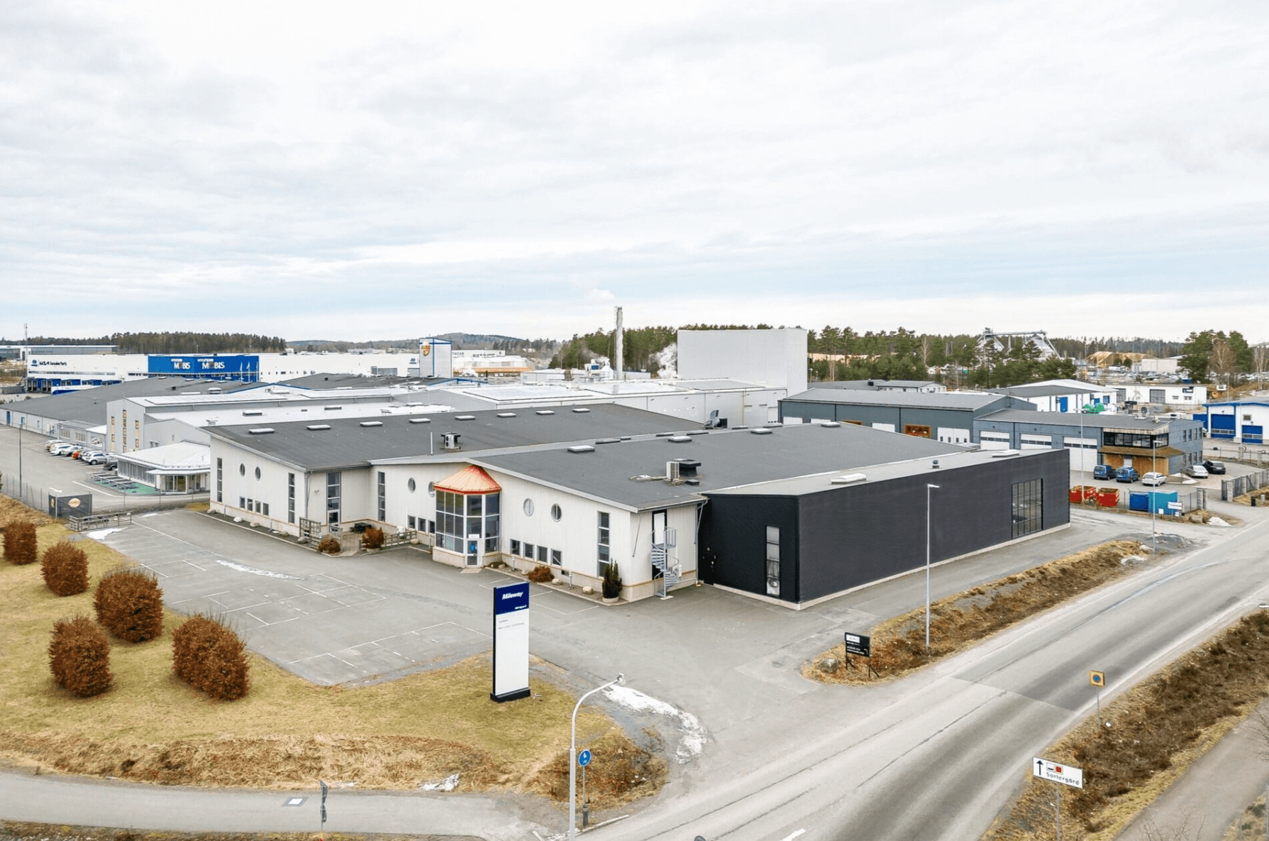 Aerial view of an industrial facility with multiple buildings, parking areas, and surrounding roads in an industrial estate on a cloudy day.