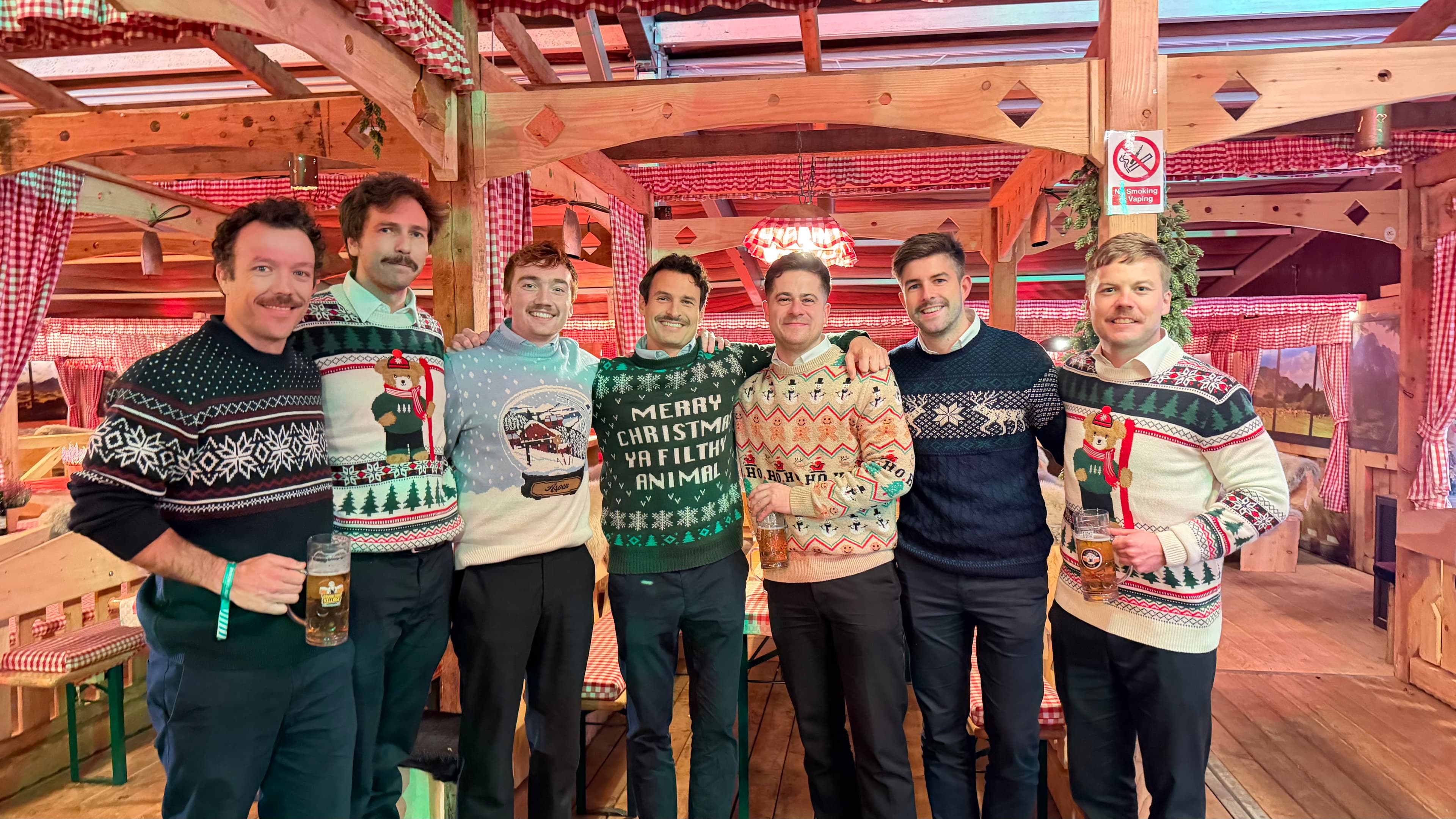 Seven men pose together indoors, all wearing festive Christmas jumpers. Two hold beer tankards, and the setting features wooden beams and checked decorations.