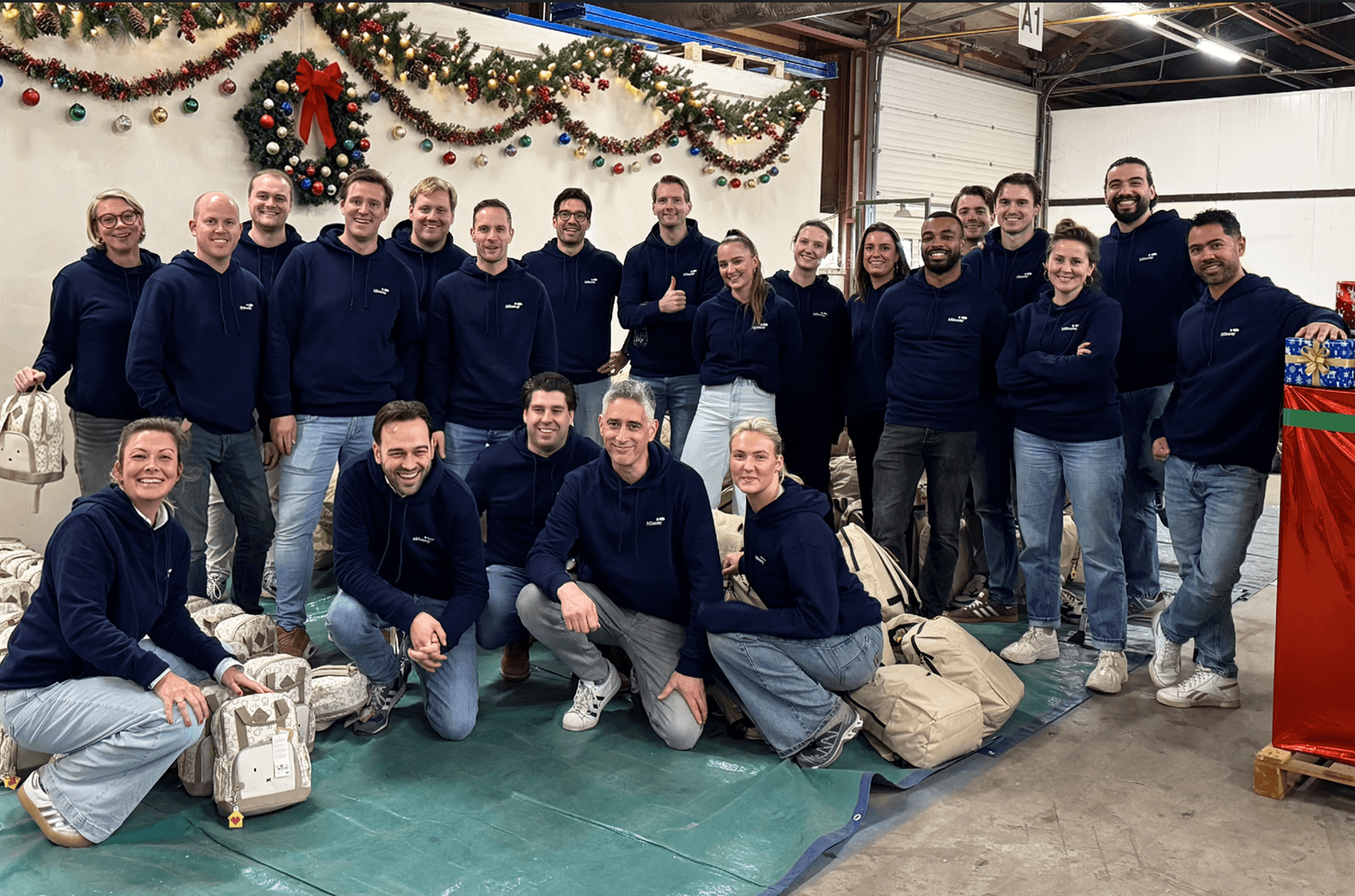 A group of adults in matching navy blue sweatshirts pose for a photo inside a decorated warehouse, surrounded by large bags and festive holiday decorations.
