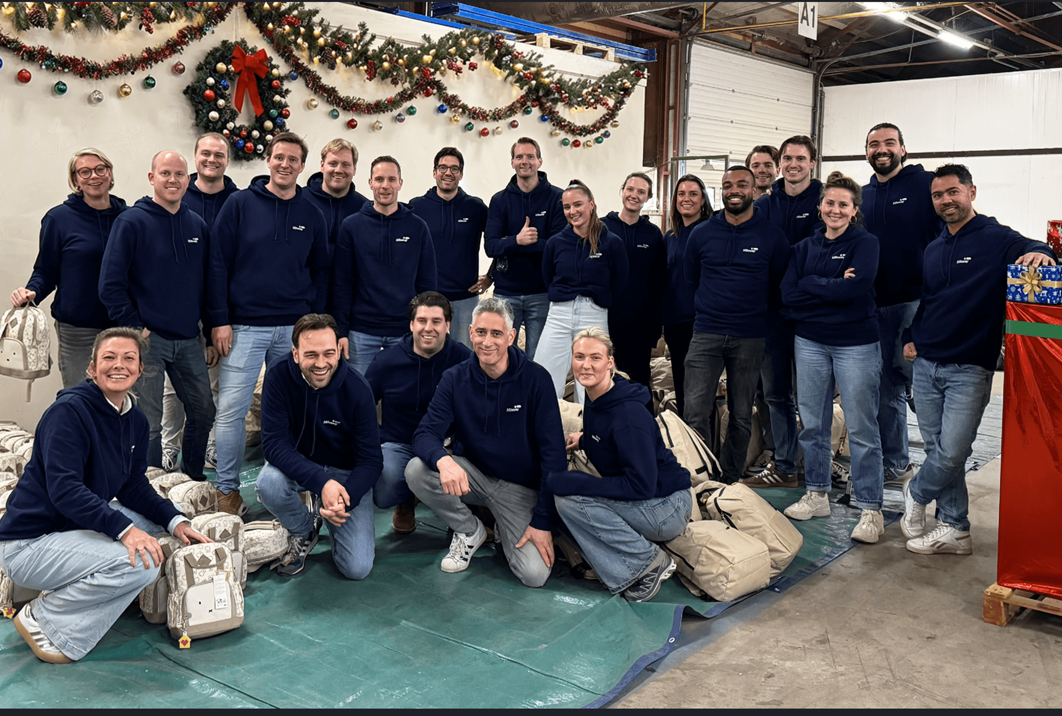 A group of adults in matching navy blue sweatshirts pose for a photo inside a decorated warehouse, surrounded by large bags and festive holiday decorations.