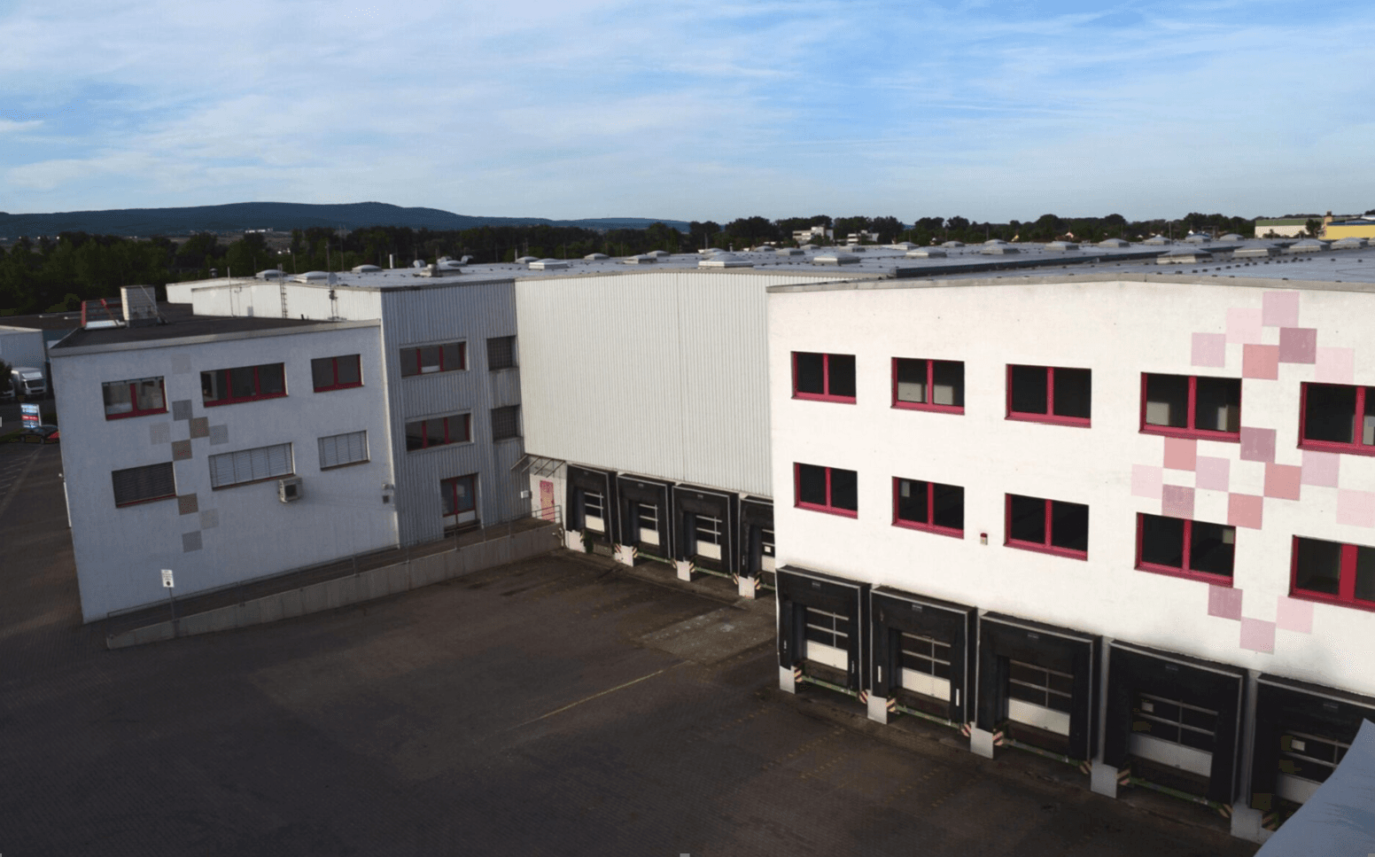 Two large, white industrial buildings with pink window frames and geometric designs, separated by a tarmacked yard under a partly cloudy sky.