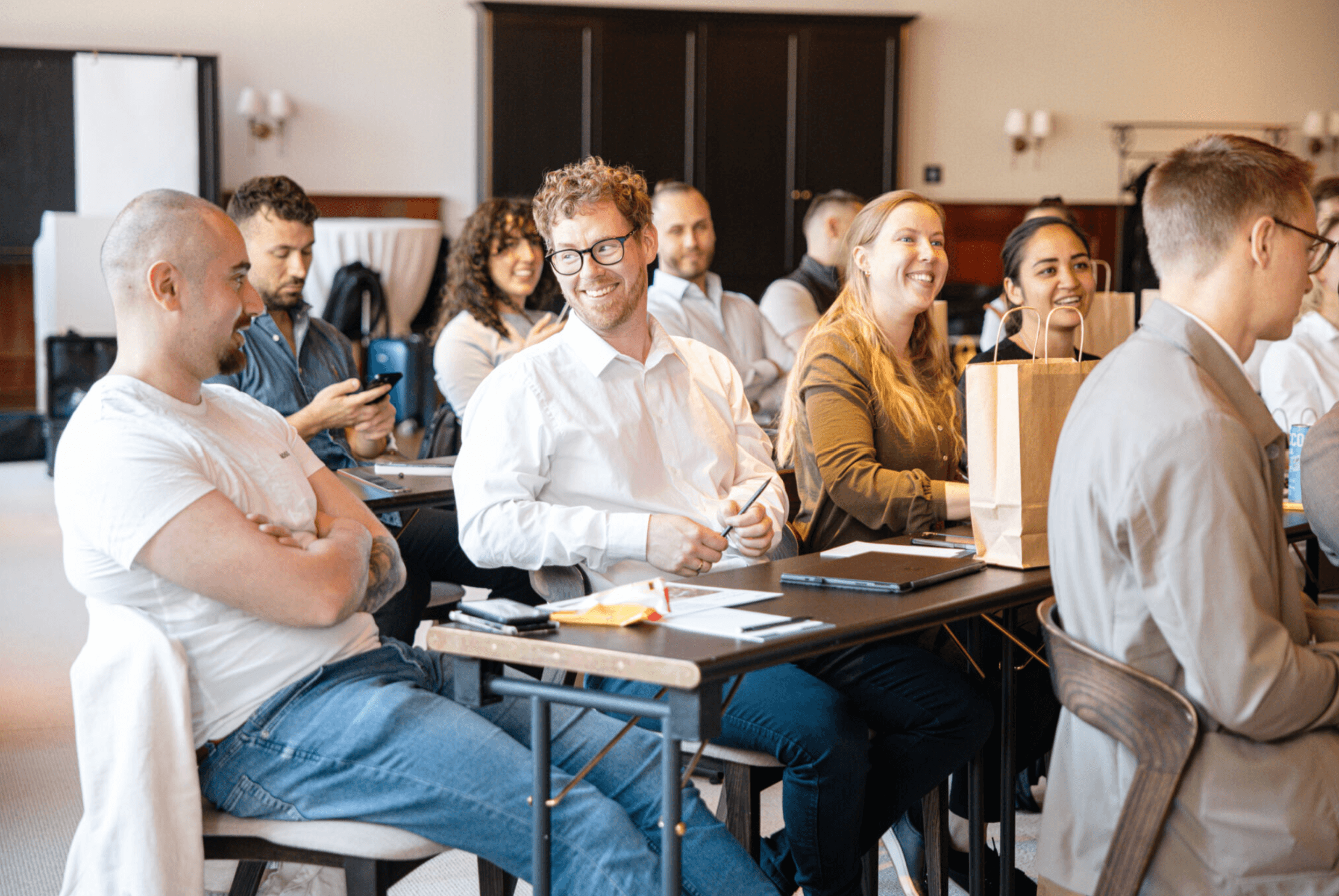 Un groupe de personnes assises à des tables dans une pièce lumineuse, écoutant attentivement et souriant lors d'une réunion ou d'un atelier.